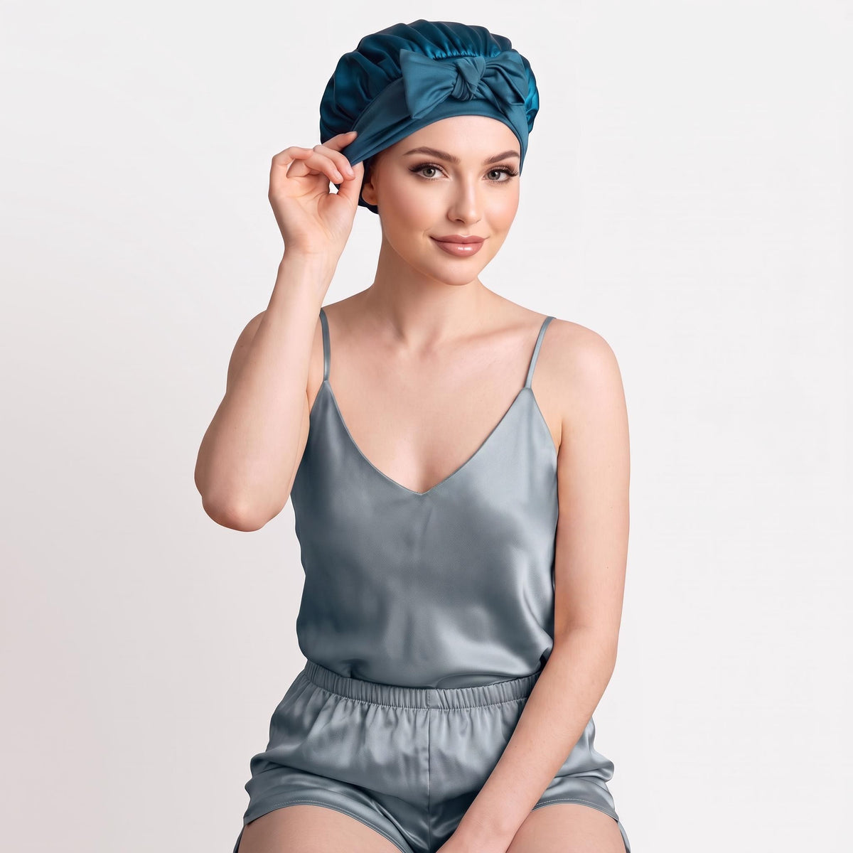 Woman wearing a teal mulberry silk sleep bonnet with a decorative bow tie, matching her satin pajama set.