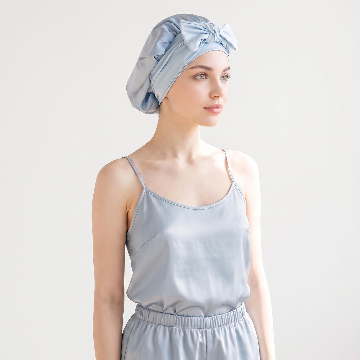 Light blue mulberry silk sleep bonnet with a decorative front bow worn by a woman in matching silk pajamas.