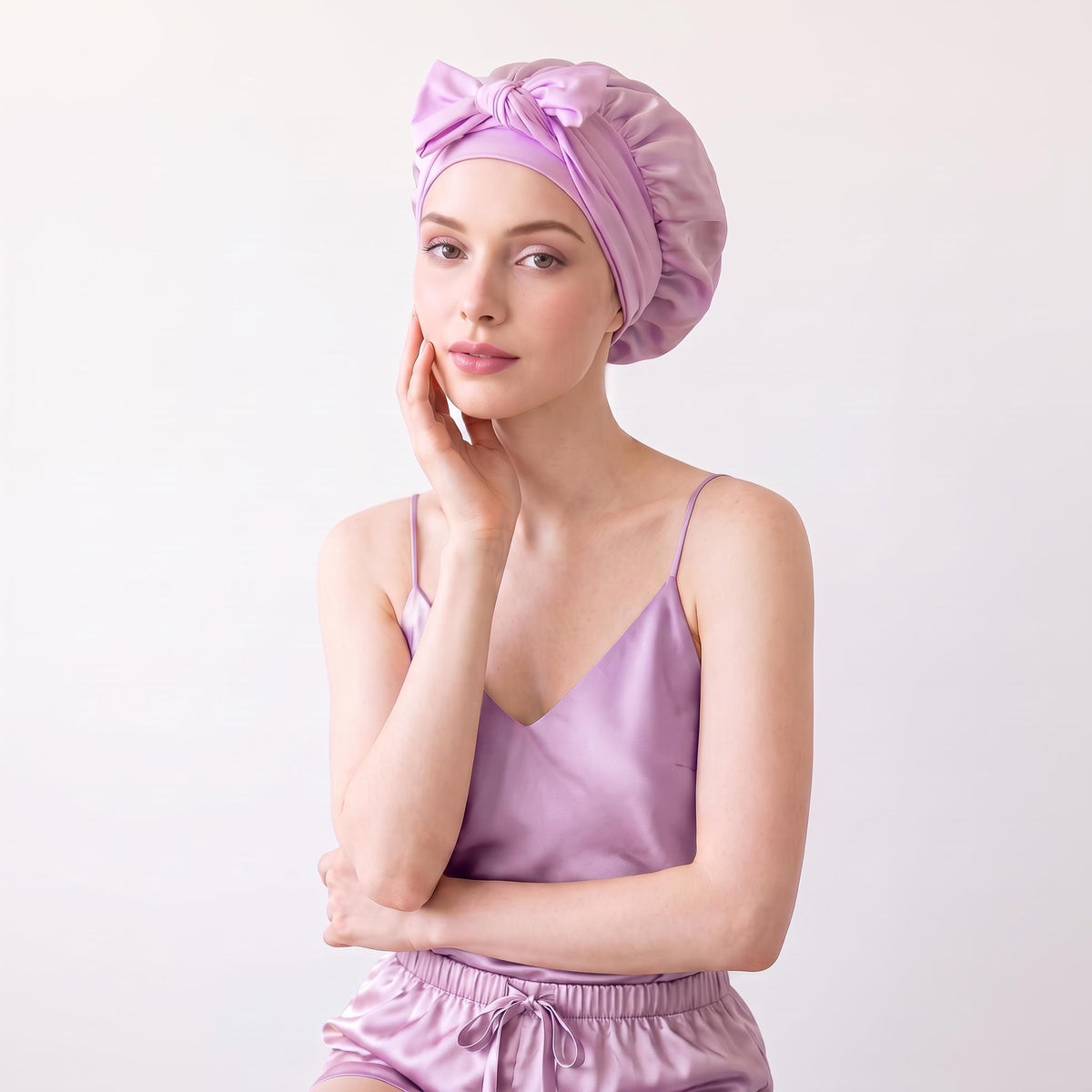 Woman wearing a light purple mulberry silk sleep bonnet with a decorative front bow tie, matching her satin pajama set.