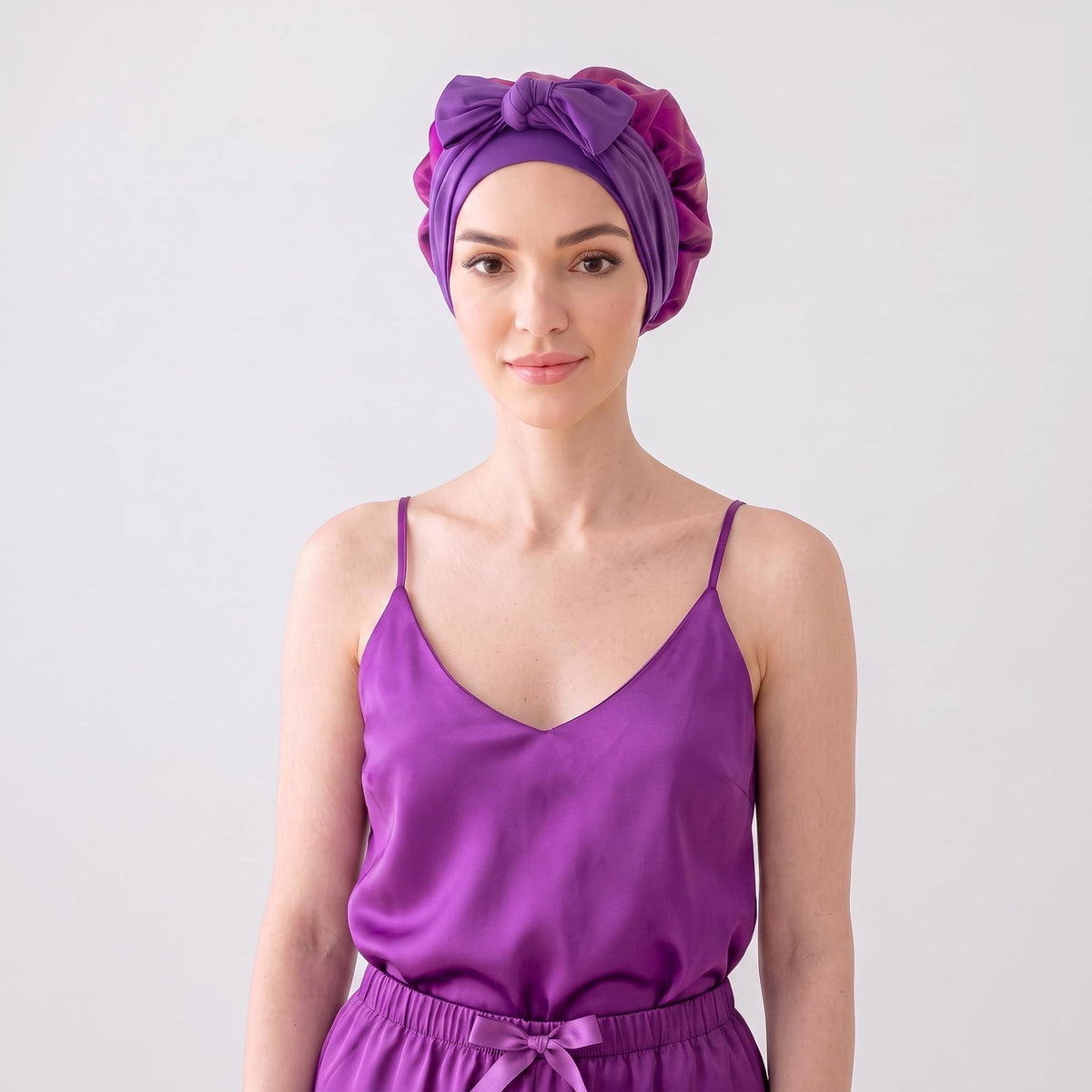 Woman wearing a purple mulberry silk sleep bonnet with an elegant front bow tie against a light background.