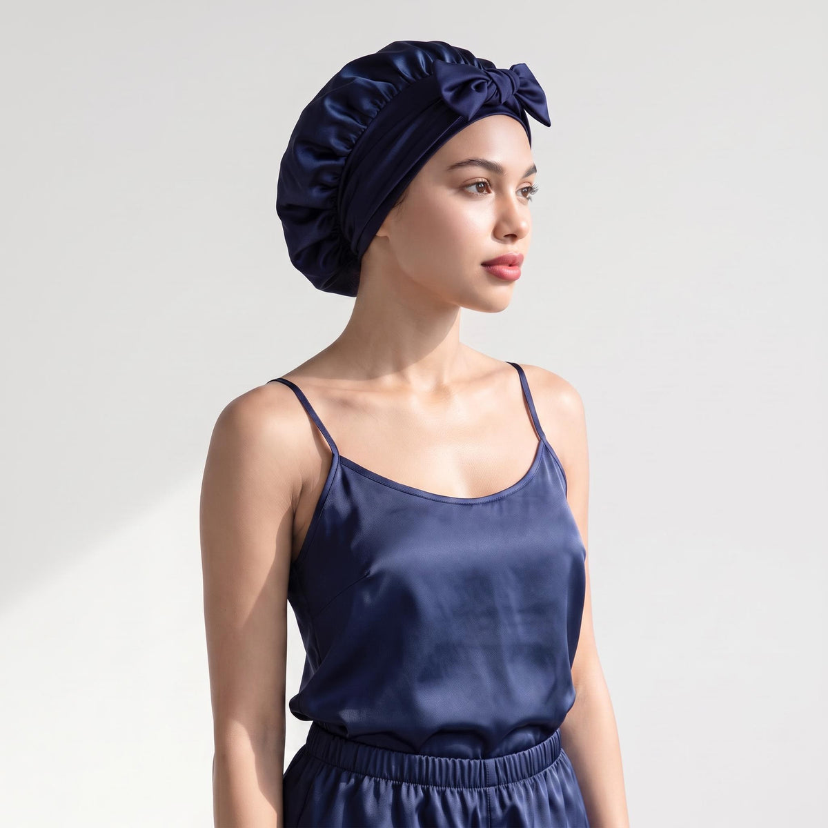 Navy blue mulberry silk sleep bonnet with a decorative bow tie worn by a woman in matching silk pajamas.