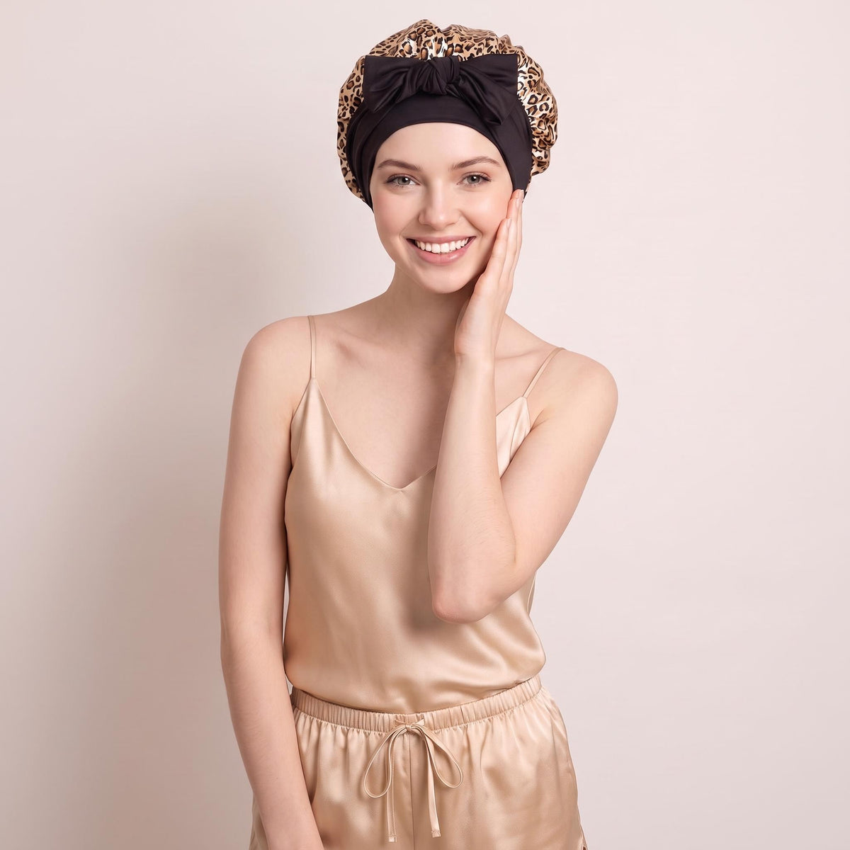 Woman wearing a leopard print mulberry silk sleep bonnet with a black bow tie, paired with matching champagne silk pajamas.