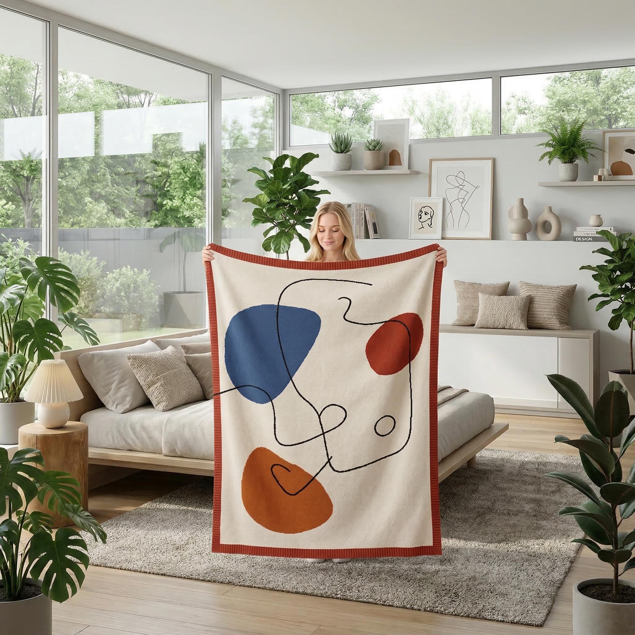 Modern abstract knitted throw blanket in earthy tones with organic shapes, held by a woman in a bright living room.