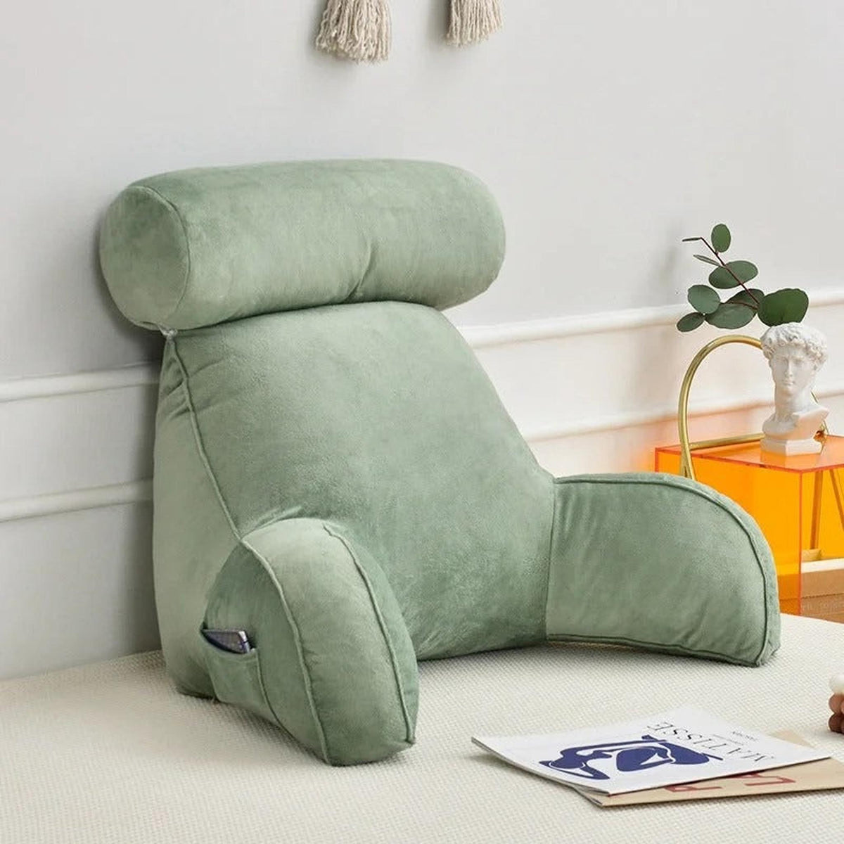 Adjustable sage green velour backrest pillow with arms, headrest, and side pocket on a bed.