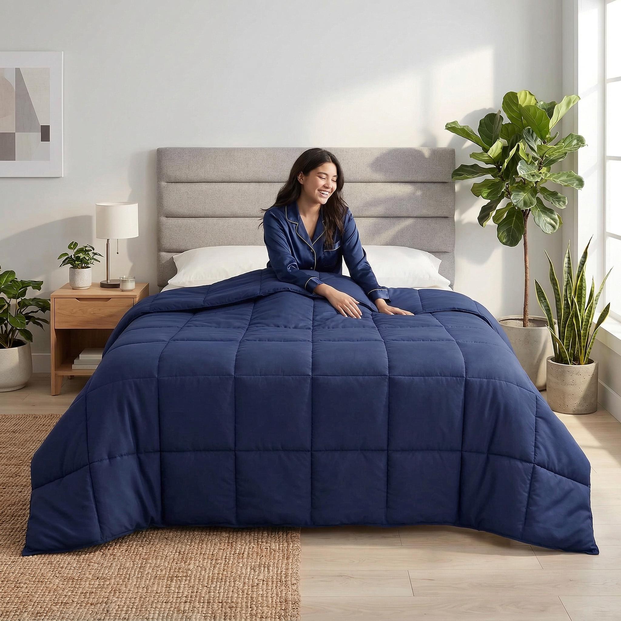 Smiling woman in blue pajamas sitting on a bed with a navy blue box-stitched all-season comforter.