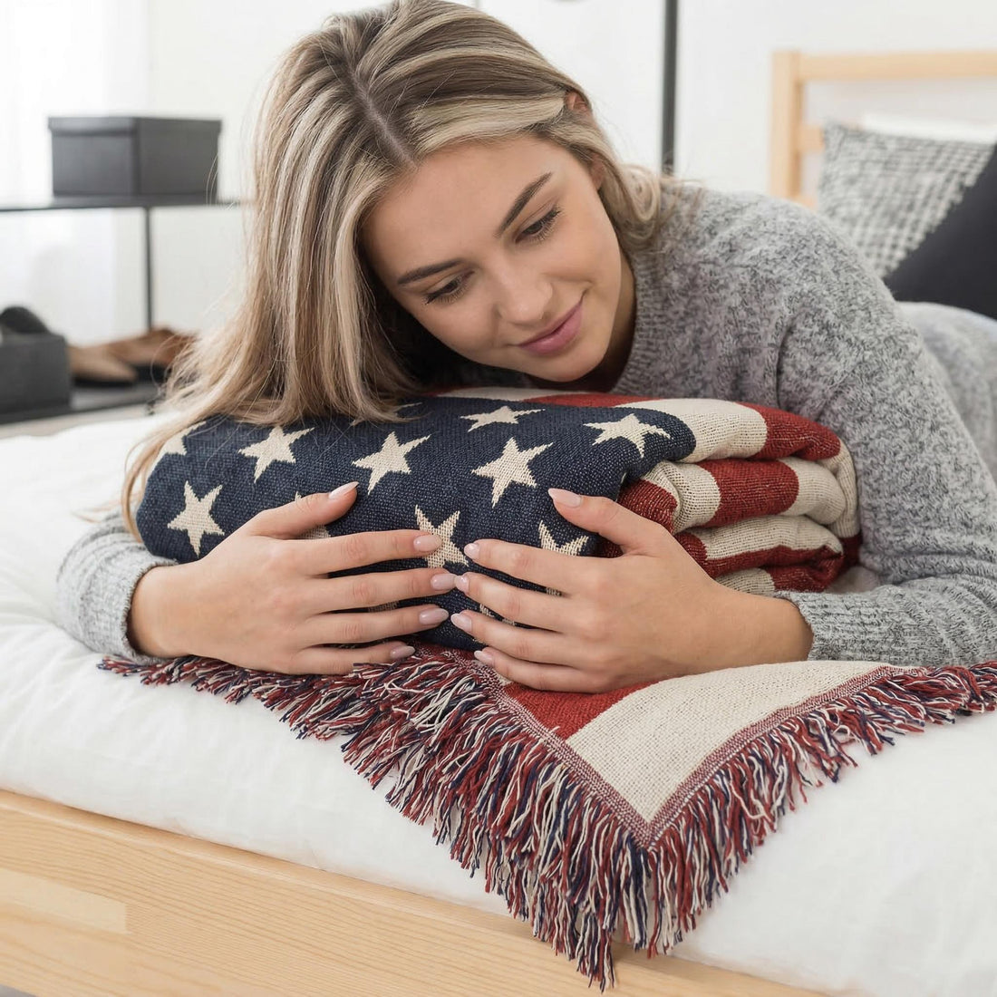 Woman relaxing on a grey chaise lounge, covered by a knitted American flag throw blanket, in a bright, modern living room.