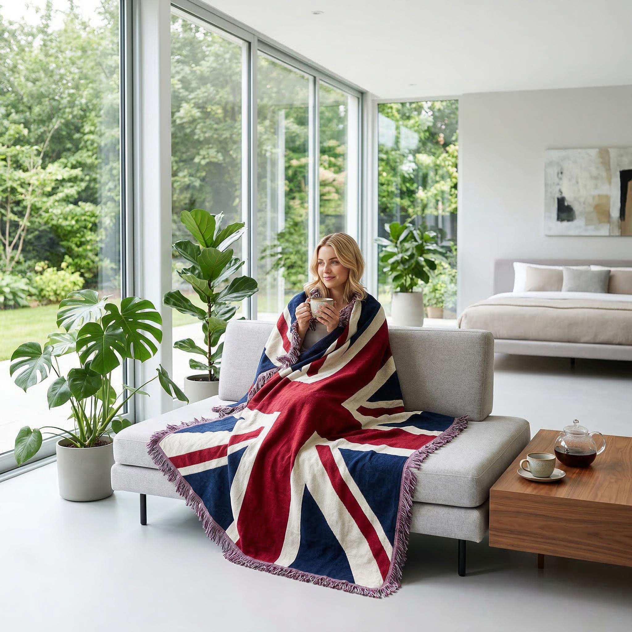 Woman on a couch wrapped in a knitted Union Jack throw blanket with fringe, holding a mug.