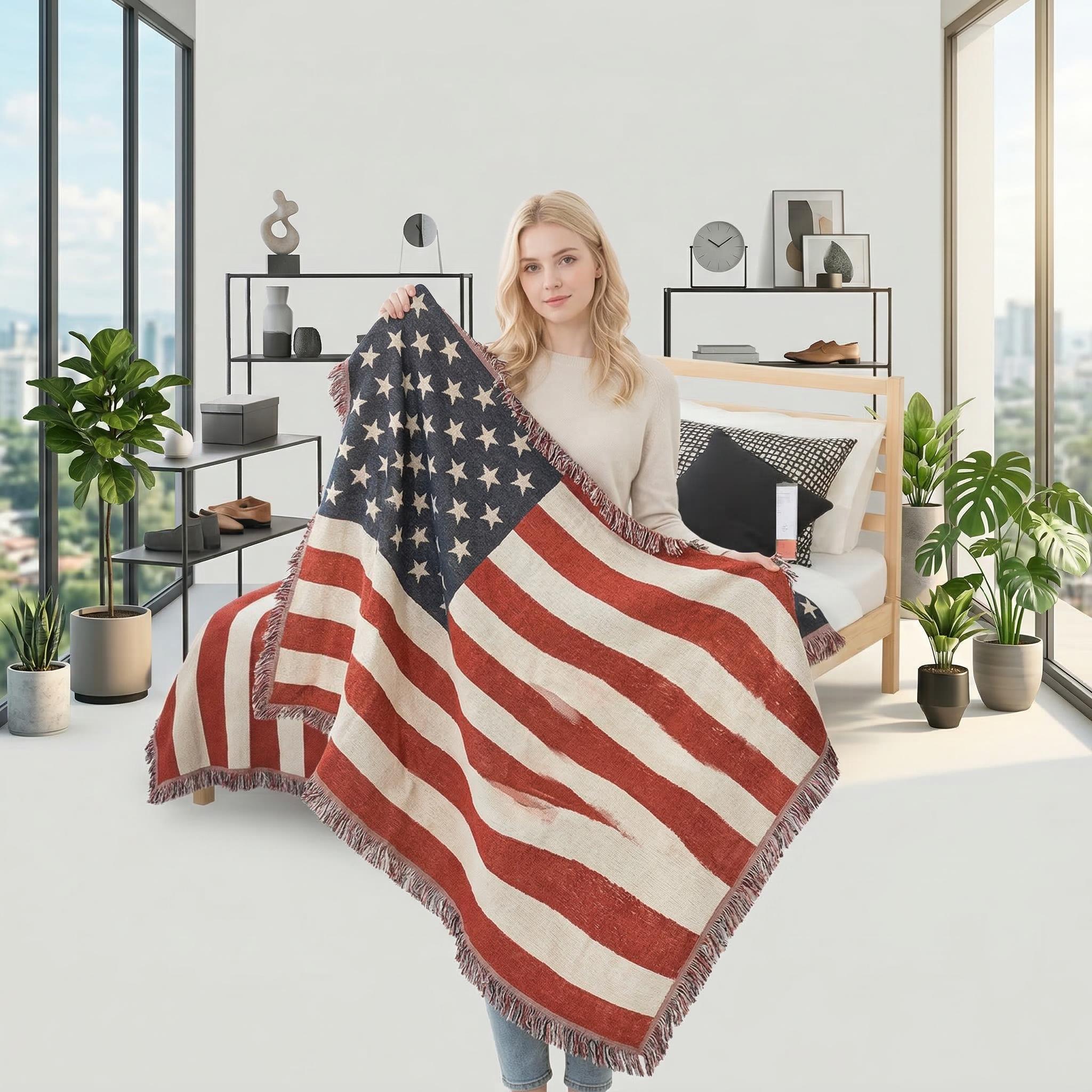 Blonde woman holding a large, distressed American flag knitted throw blanket with fringe in a modern bedroom.
