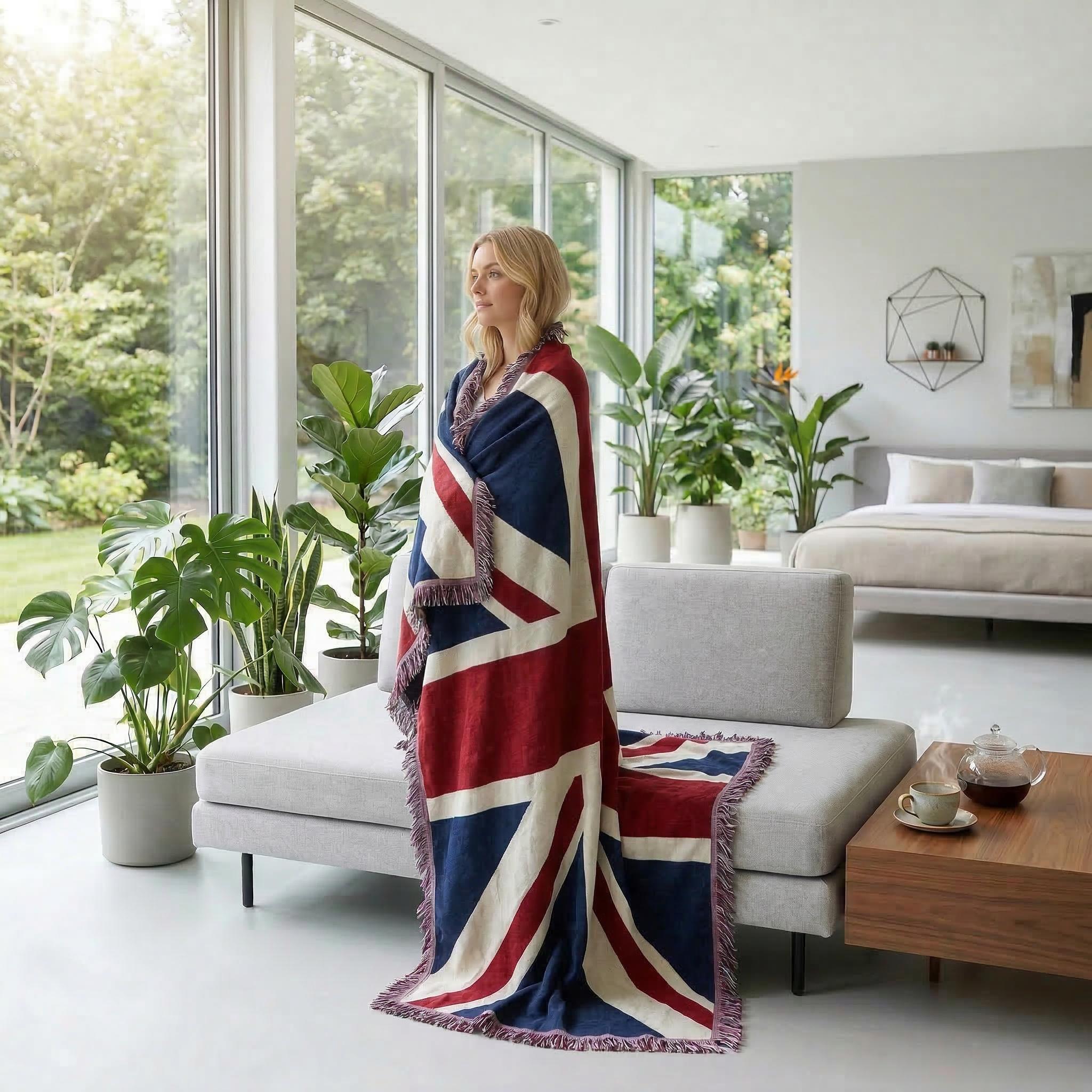 Blonde woman wrapped in a knitted Union Jack throw blanket, standing by a window in a modern living room.