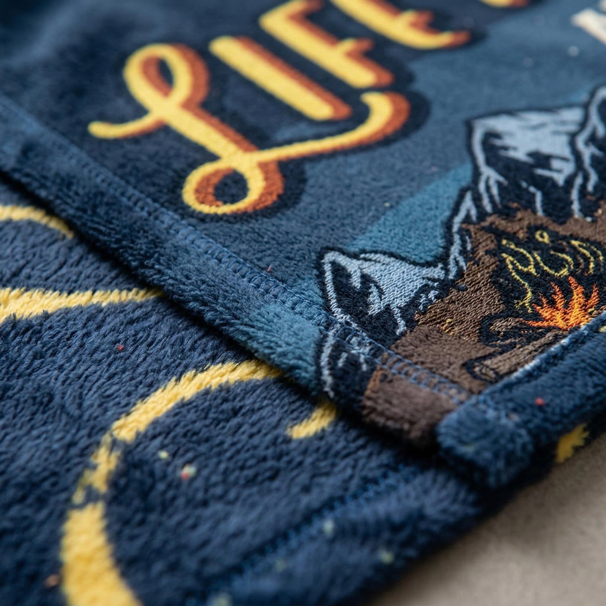 Soft navy flannel blanket with yellow script and a retro landscape design featuring mountains and a campfire.