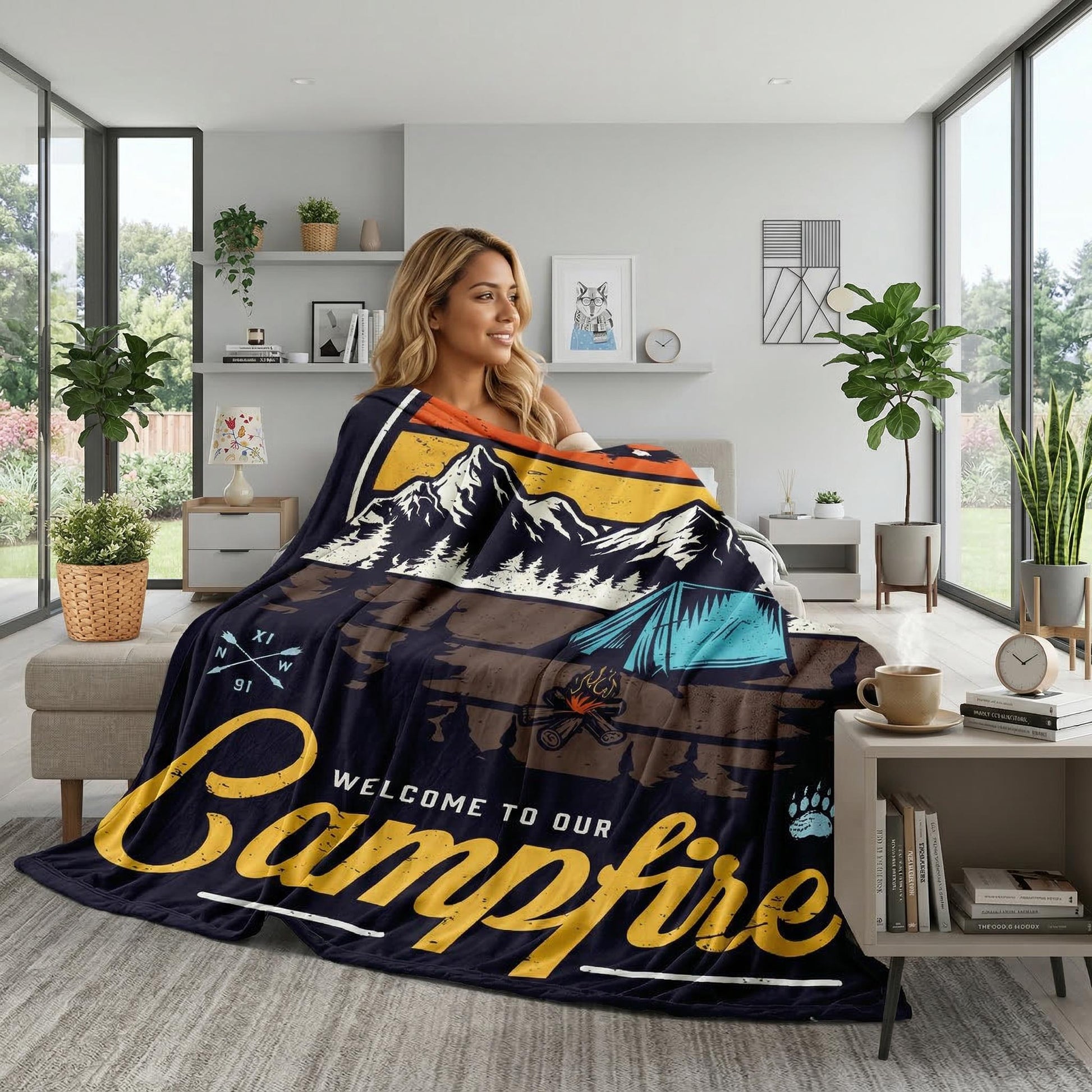 Blonde woman wrapped in a dark blue flannel blanket with a "Welcome to Our Campfire" landscape design.