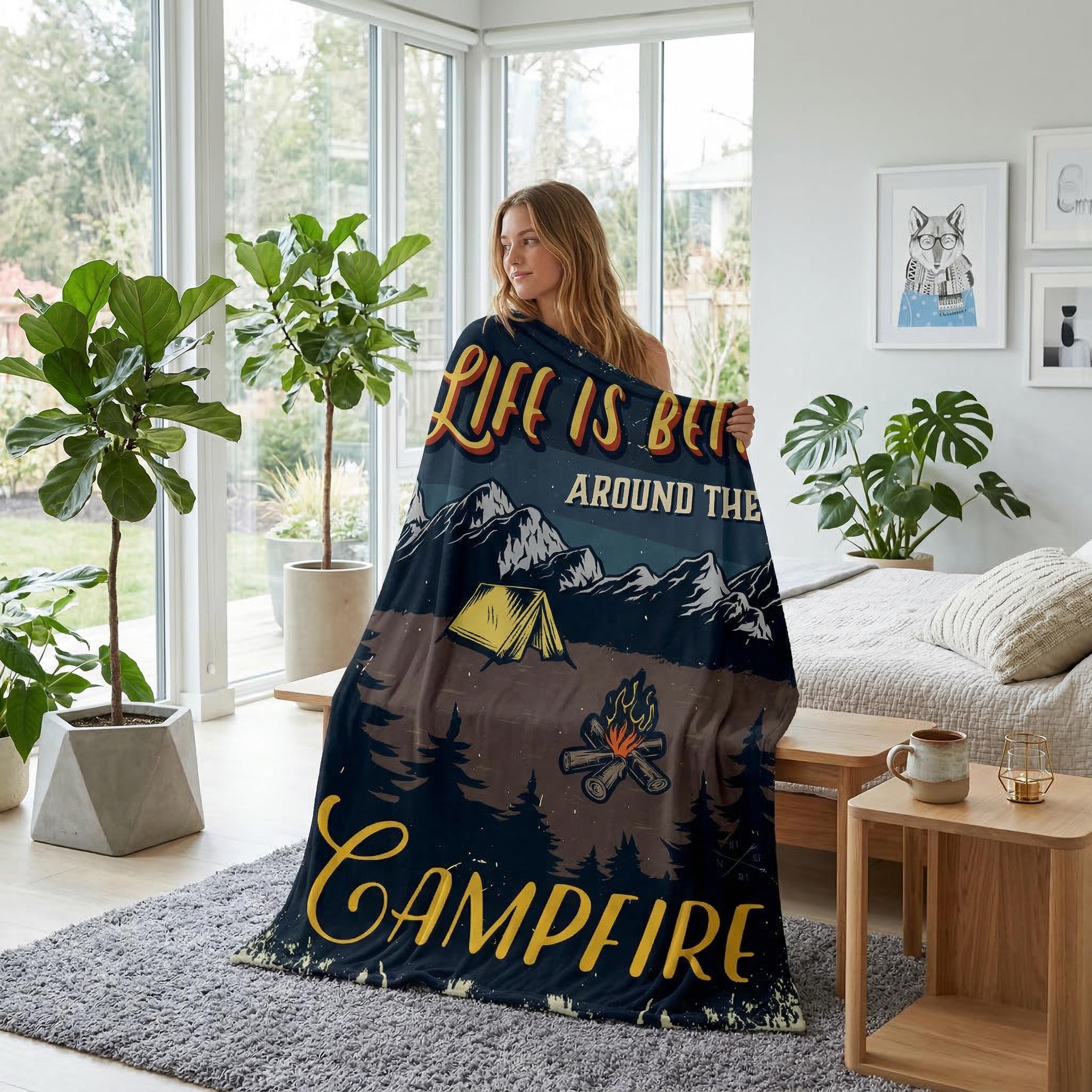 Woman wrapped in a dark blue flannel blanket with a "Life is Better Around the Campfire" landscape design.