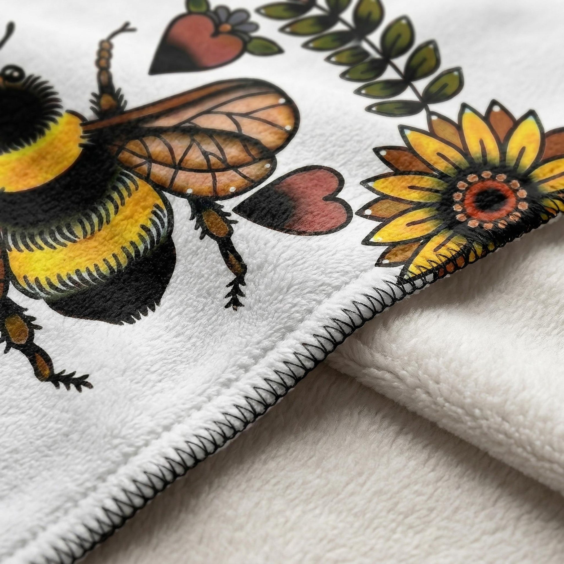 White plush blanket with a vibrant bee, sunflower, heart, and leaf floral print, featuring a black stitched edge.