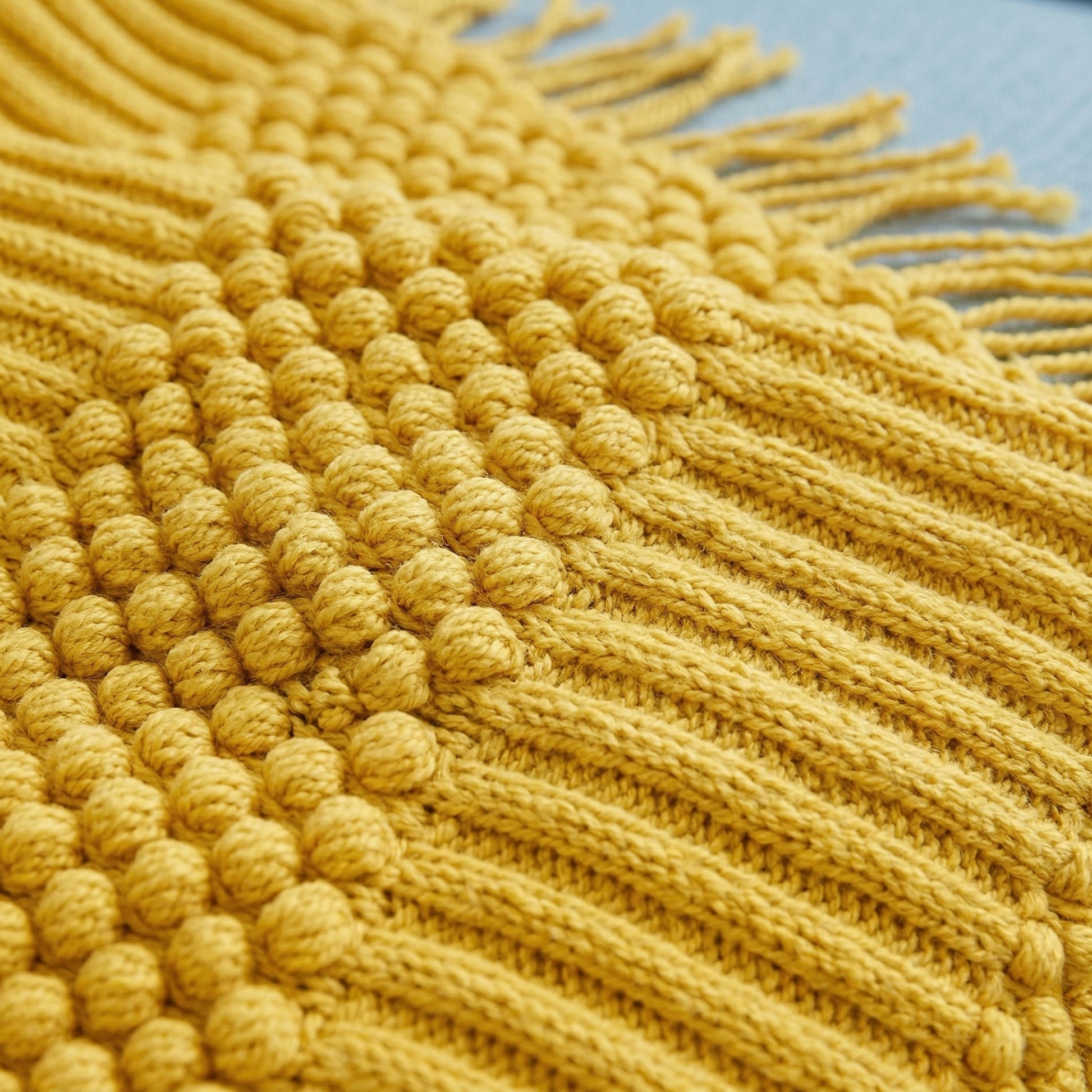 Close-up of a mustard yellow knitted acrylic blanket with textured bobble weave and tassel edges.