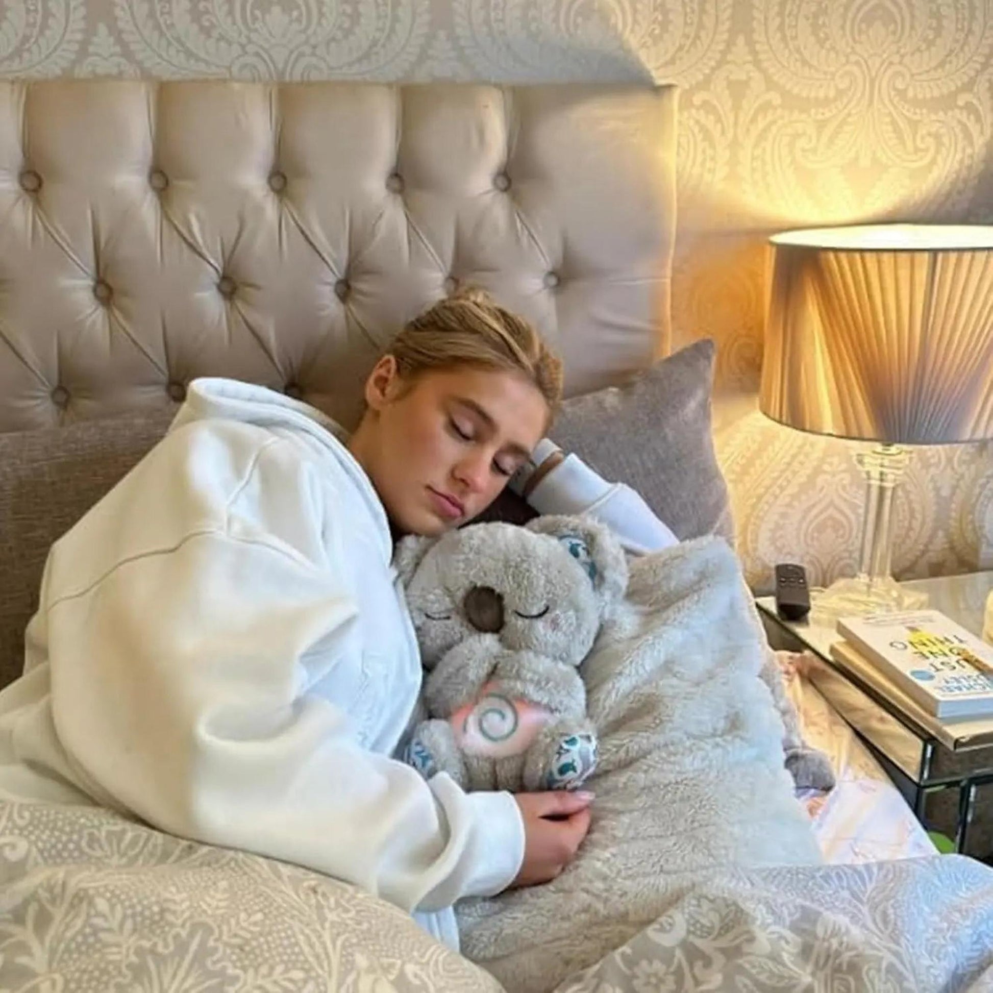 A woman sleeping in bed hugs a grey plush koala toy with a glowing belly.