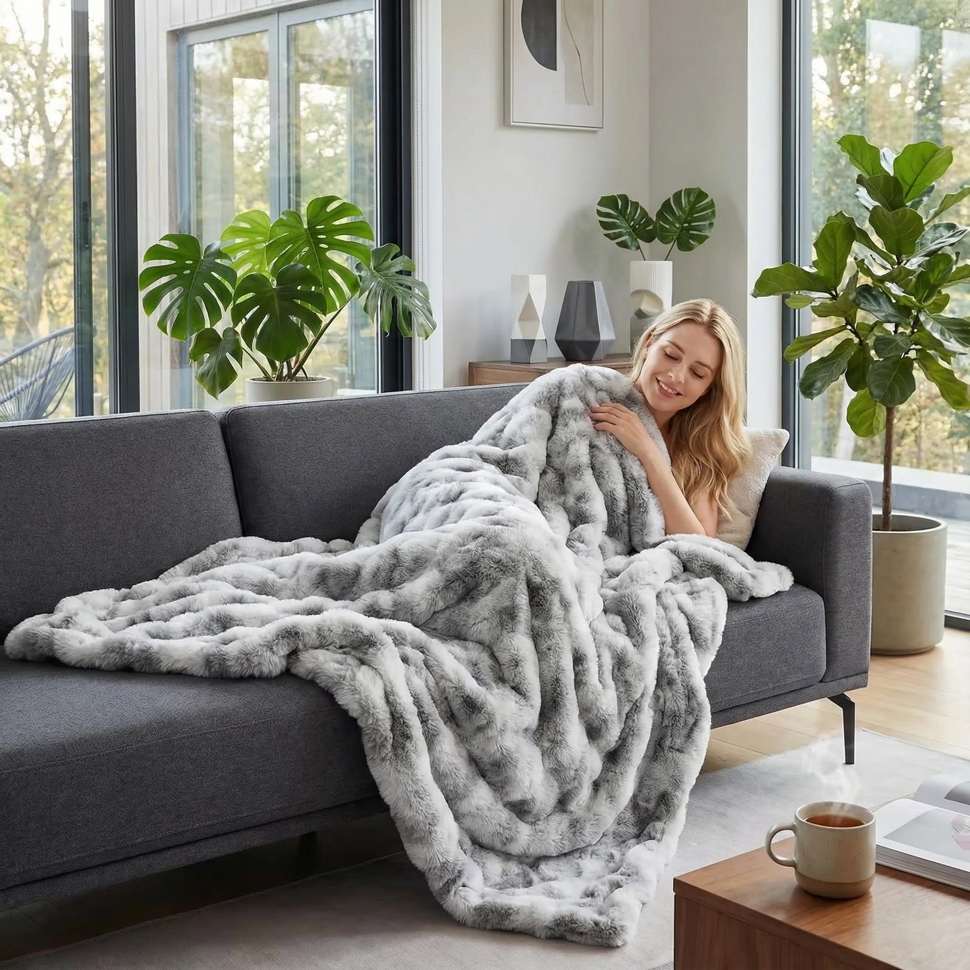 Blonde woman relaxing on a gray sofa, wrapped in a soft, gray and white bubble plush throw blanket.