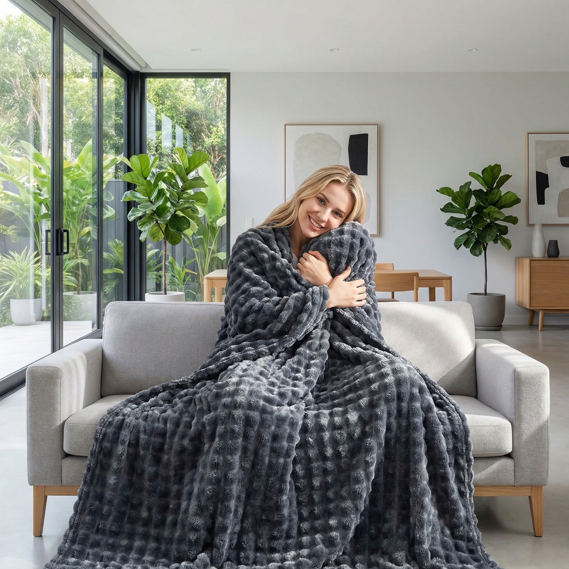 Smiling woman wrapped in a cozy, dark gray bubble rabbit flannel blanket on a light gray sofa.