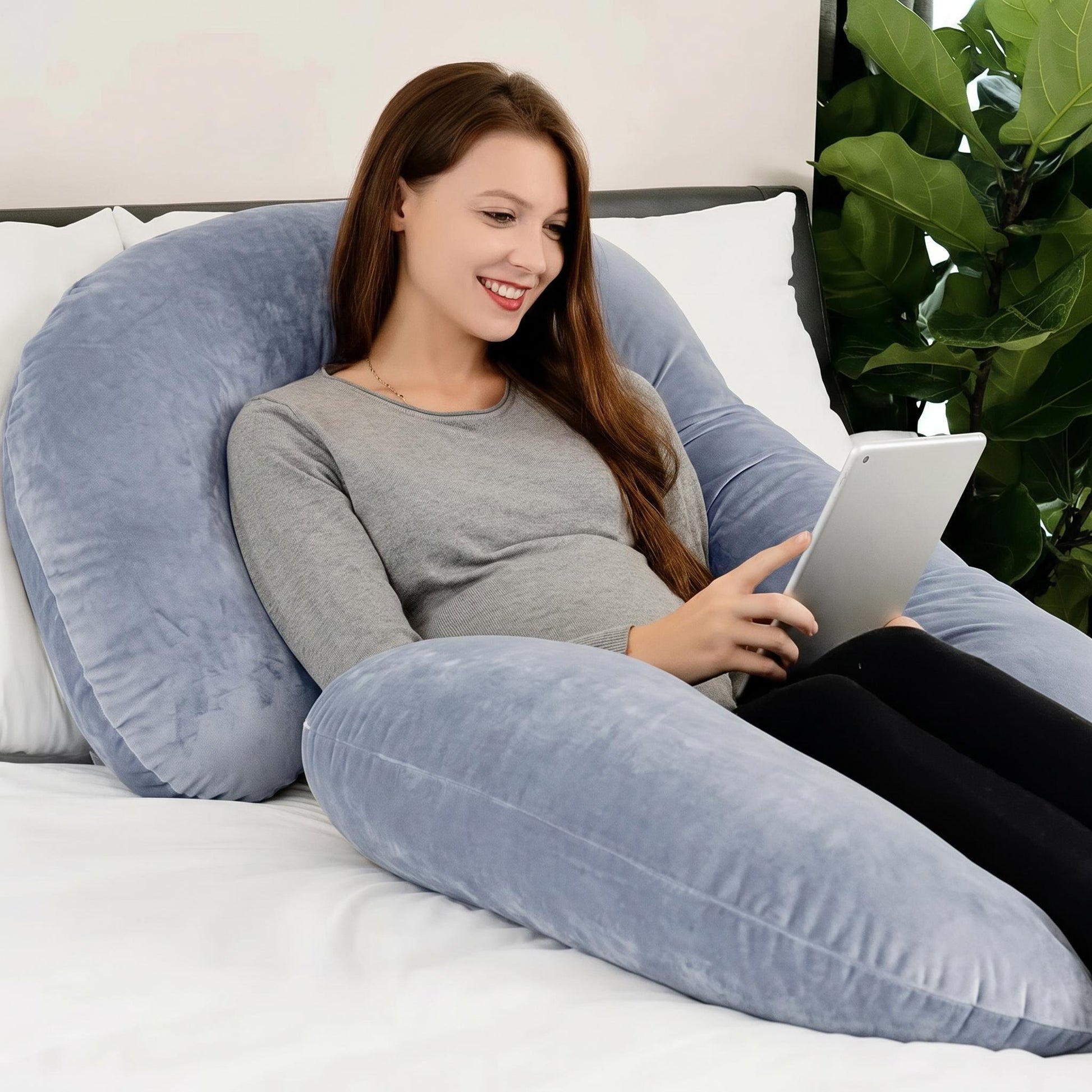 Smiling woman resting on a large, plush, light blue C-shaped support pillow, holding a tablet.