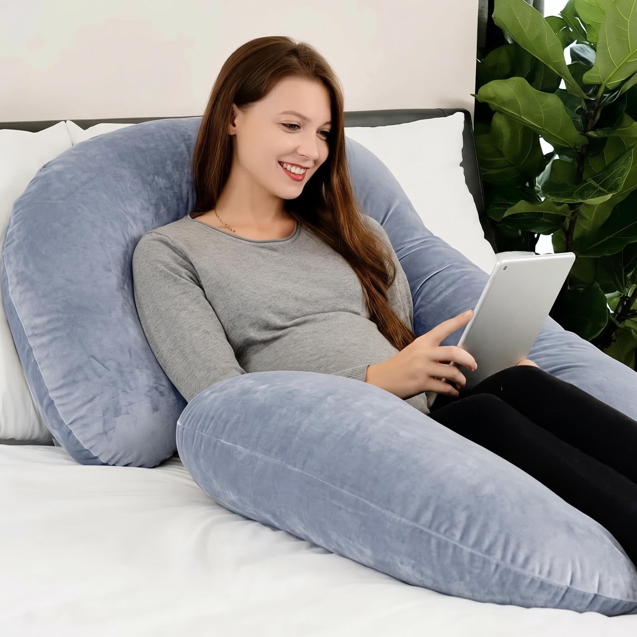 Smiling woman resting on a large, plush, light blue C-shaped support pillow, holding a tablet.