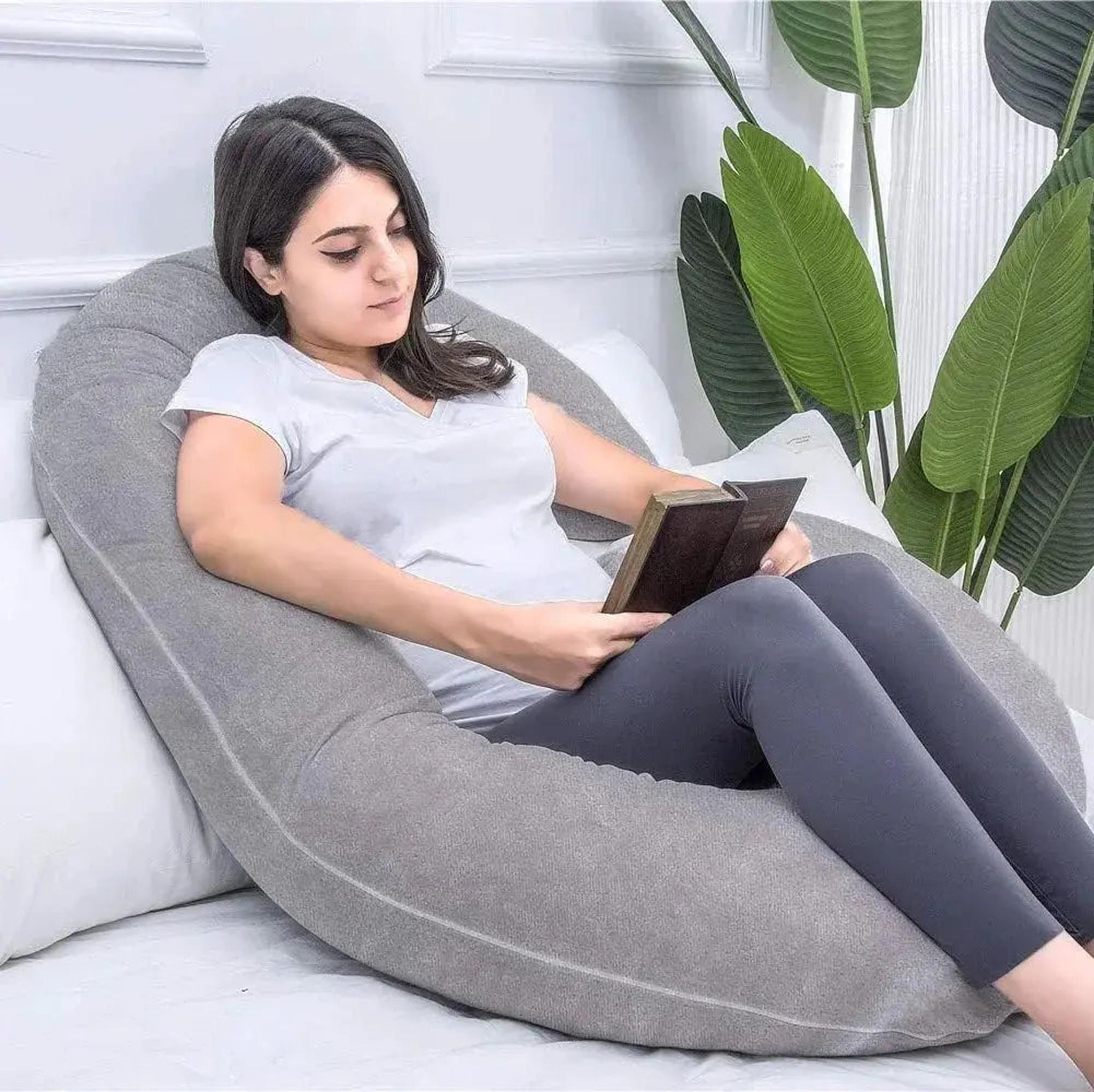 Woman relaxing with a grey C-shaped support pillow, reading a book.