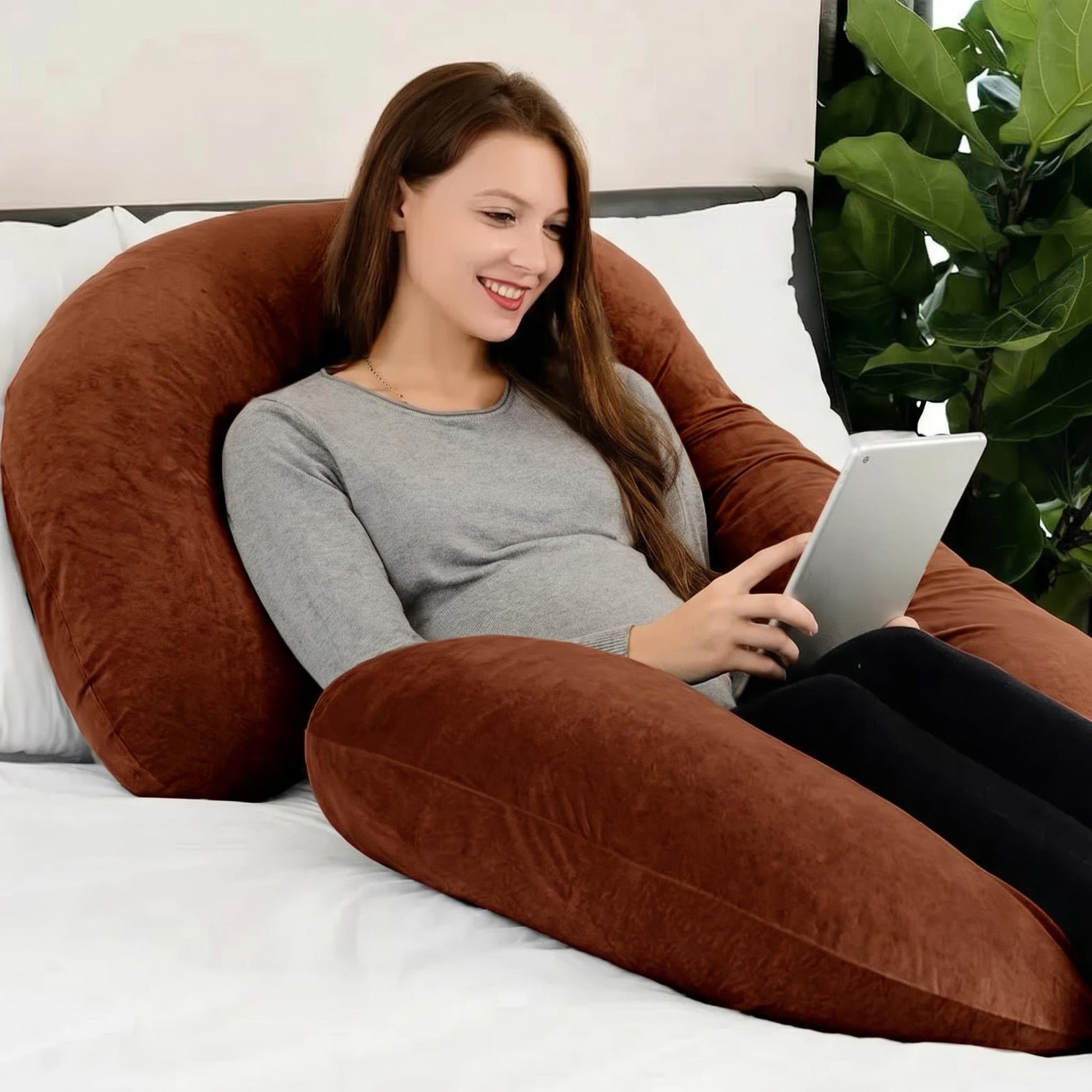 Smiling woman resting on a large brown C-shaped support pillow, using a tablet.