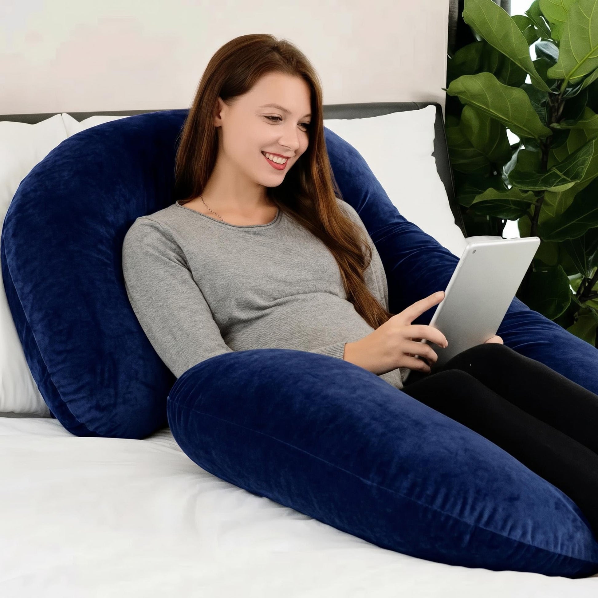 Smiling woman relaxing on a large, plush navy blue C-shaped support pillow, holding a tablet.