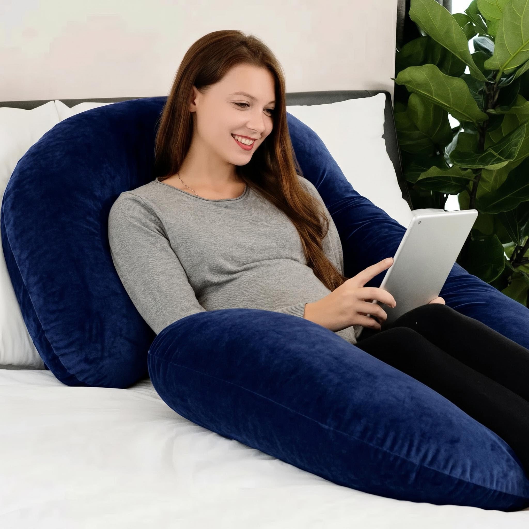 Smiling woman relaxing on a large, plush navy blue C-shaped support pillow, holding a tablet.
