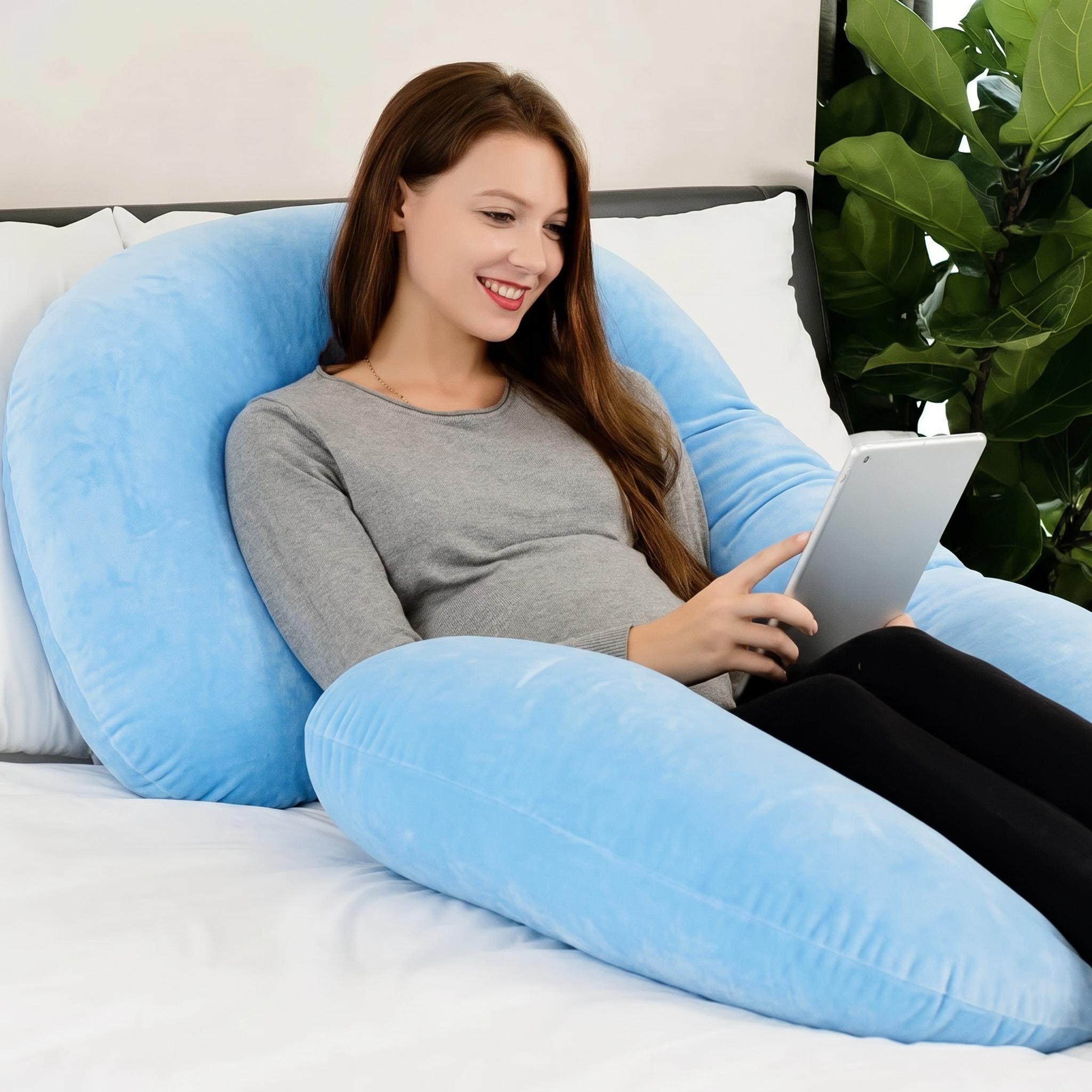 Woman relaxing in a plush blue C-shaped support pillow on a bed, holding a tablet.