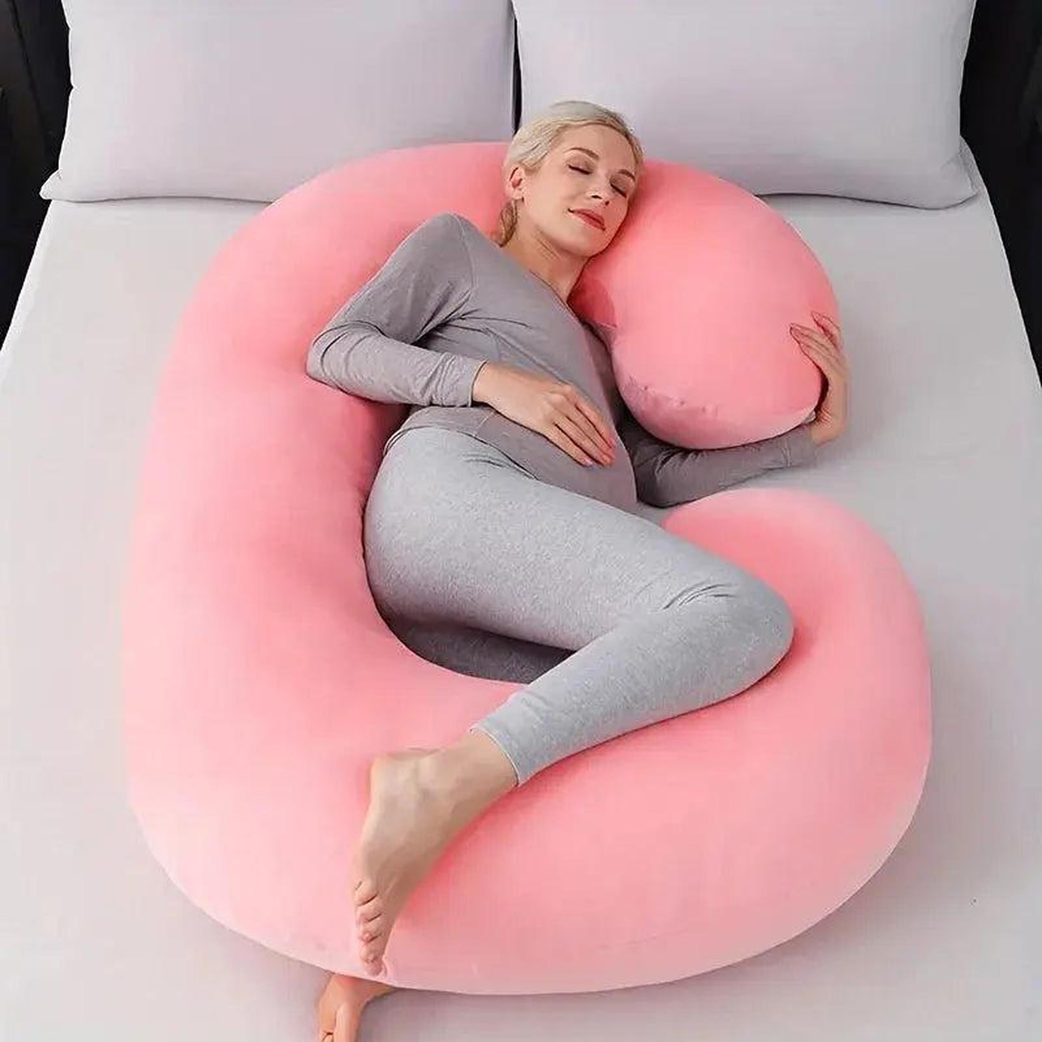 Pregnant woman resting comfortably on a large pink C-shaped support pillow on a bed.