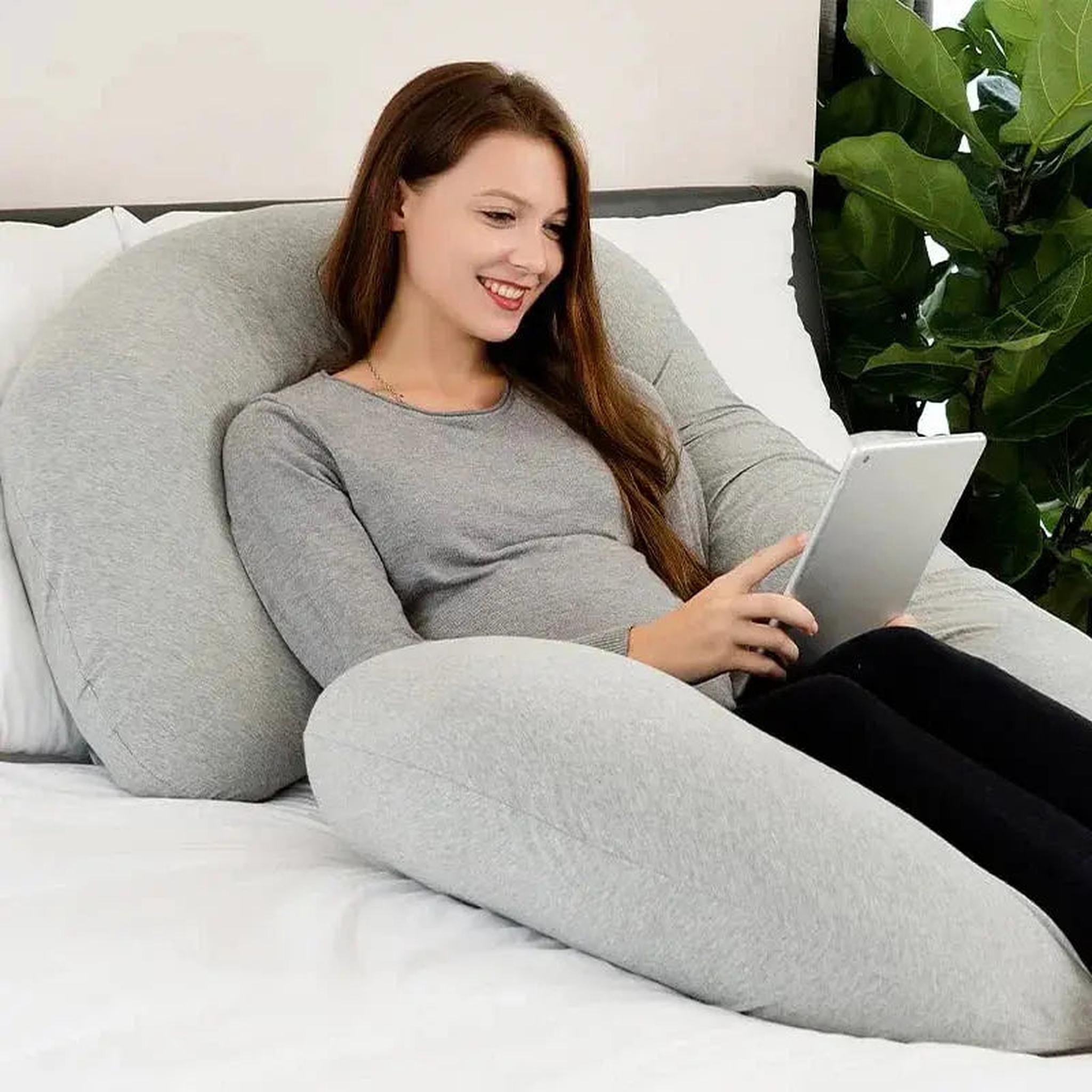 Smiling woman relaxing on a bed with a large gray C-shaped support pillow, holding a tablet.