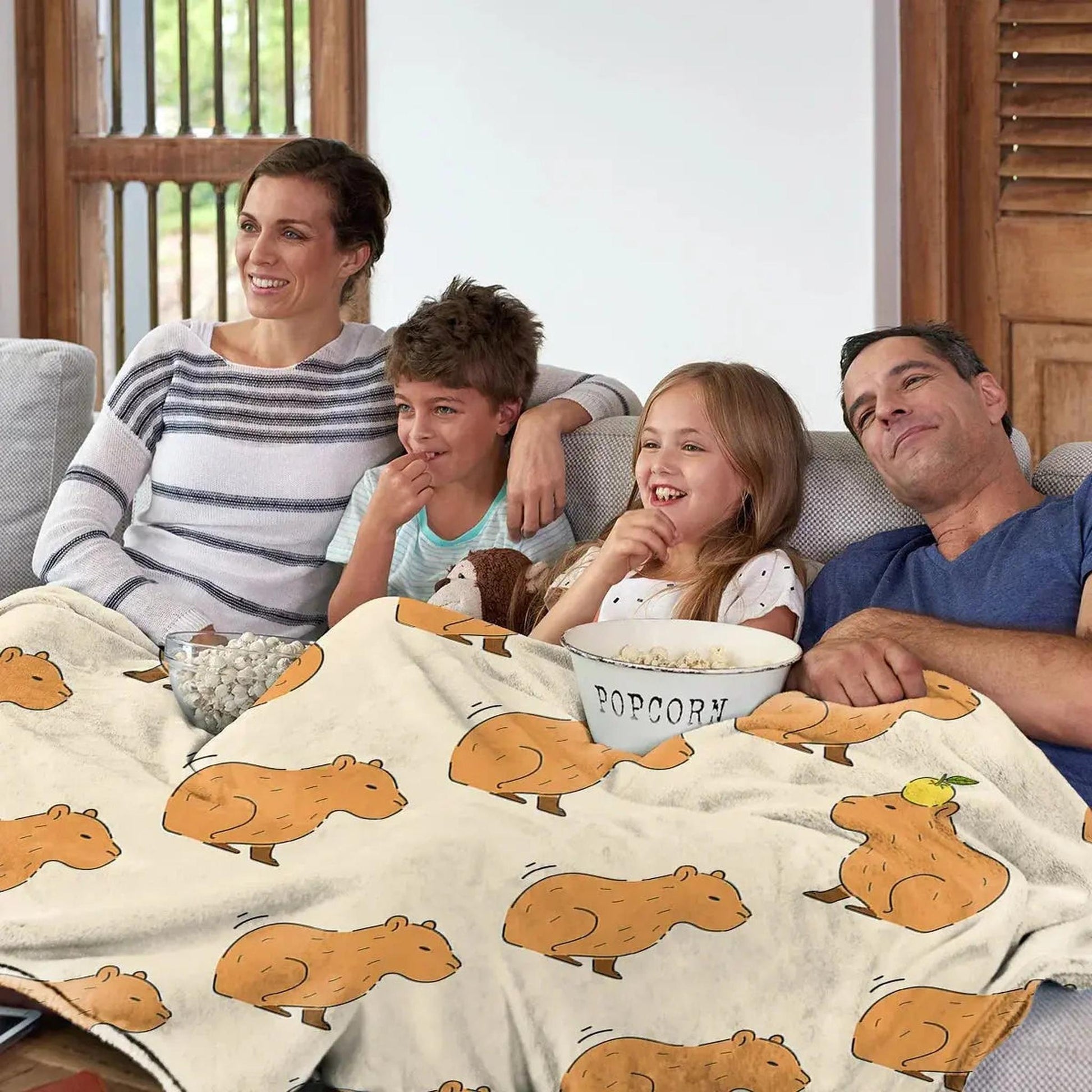 Family on a sofa enjoying a movie, covered by a cream flannel blanket with a playful capybara print.