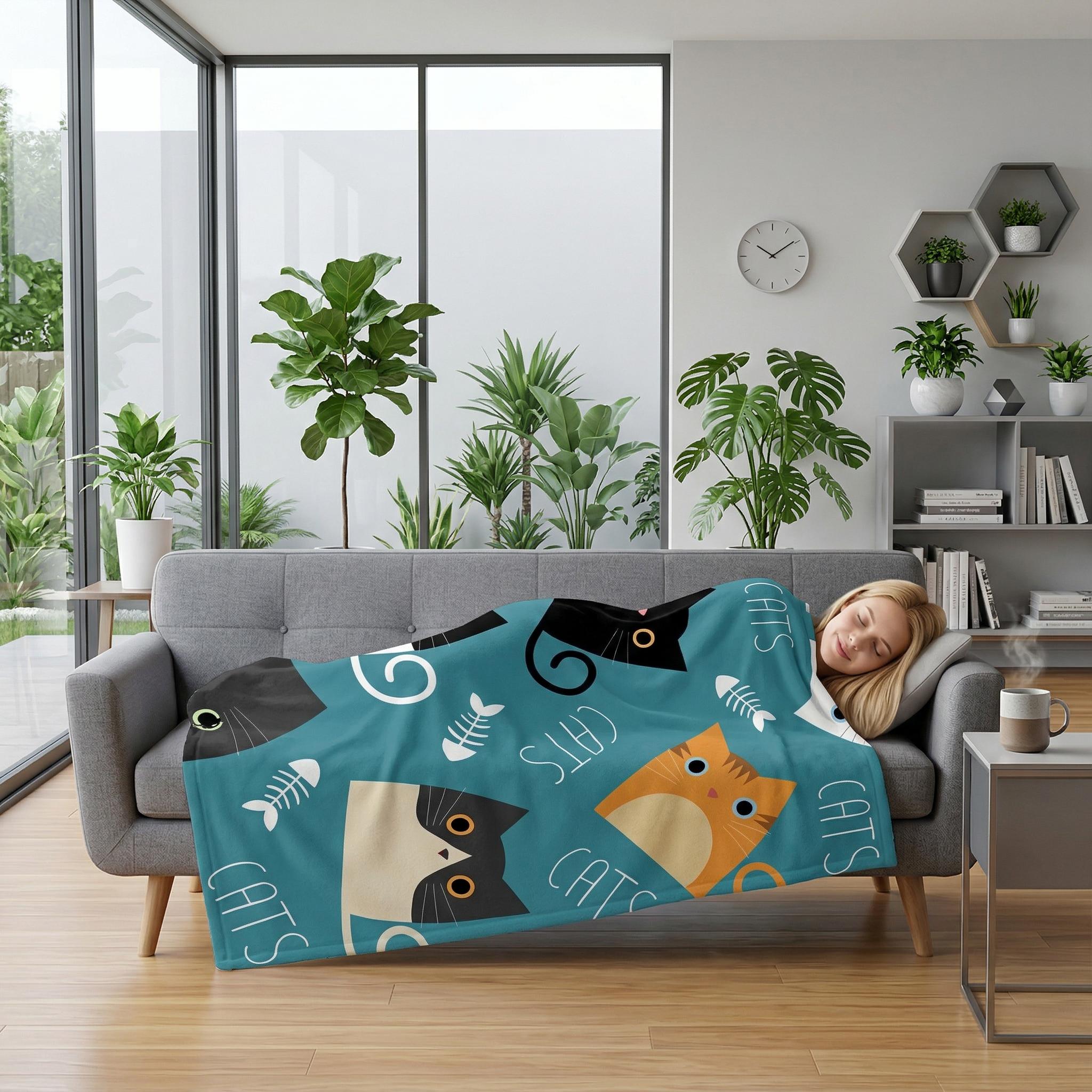 Woman sleeping on a gray sofa, covered by a teal flannel blanket with cartoon cats and fishbones.