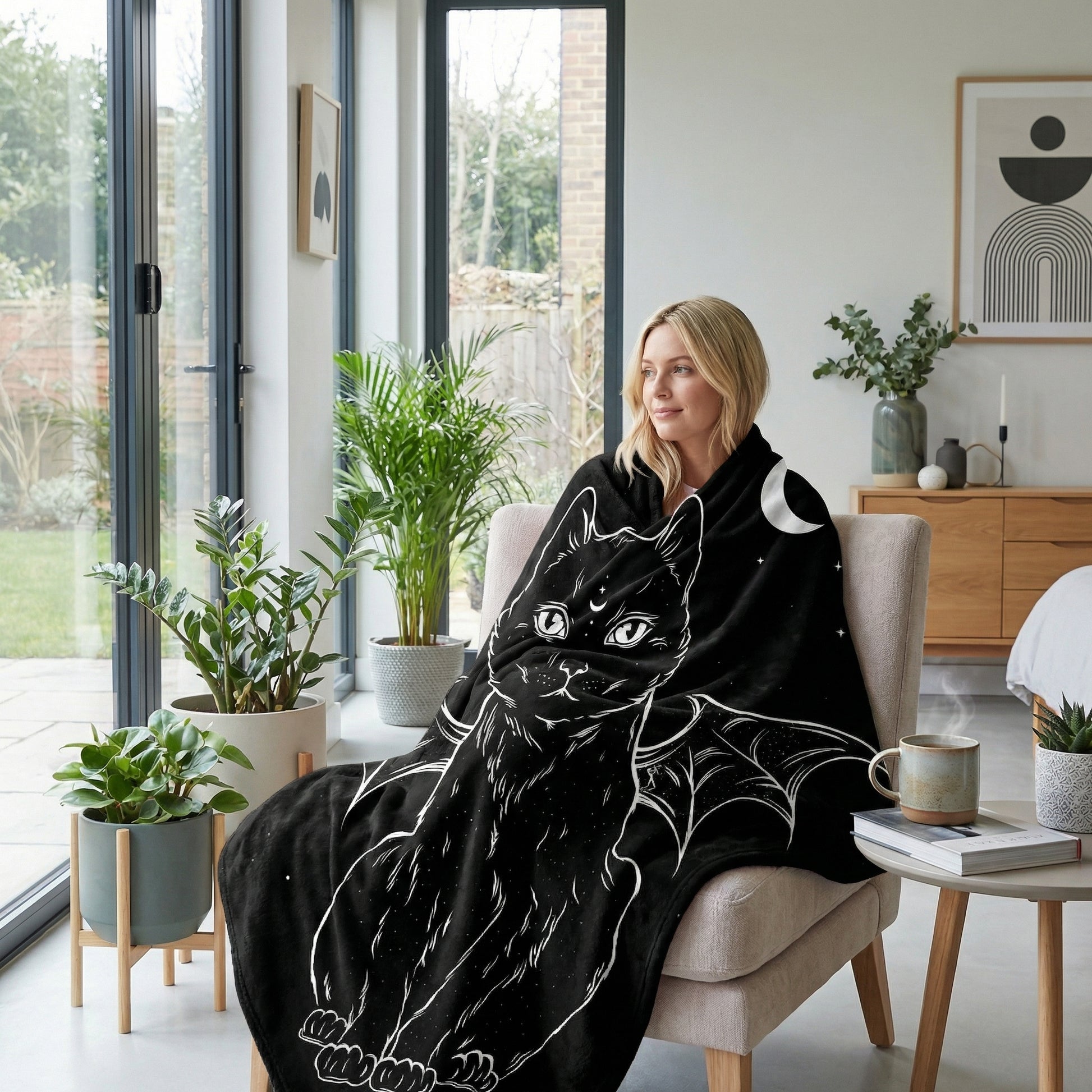 Blonde woman wrapped in a black flannel throw blanket featuring a white cat with wings and a crescent moon design.