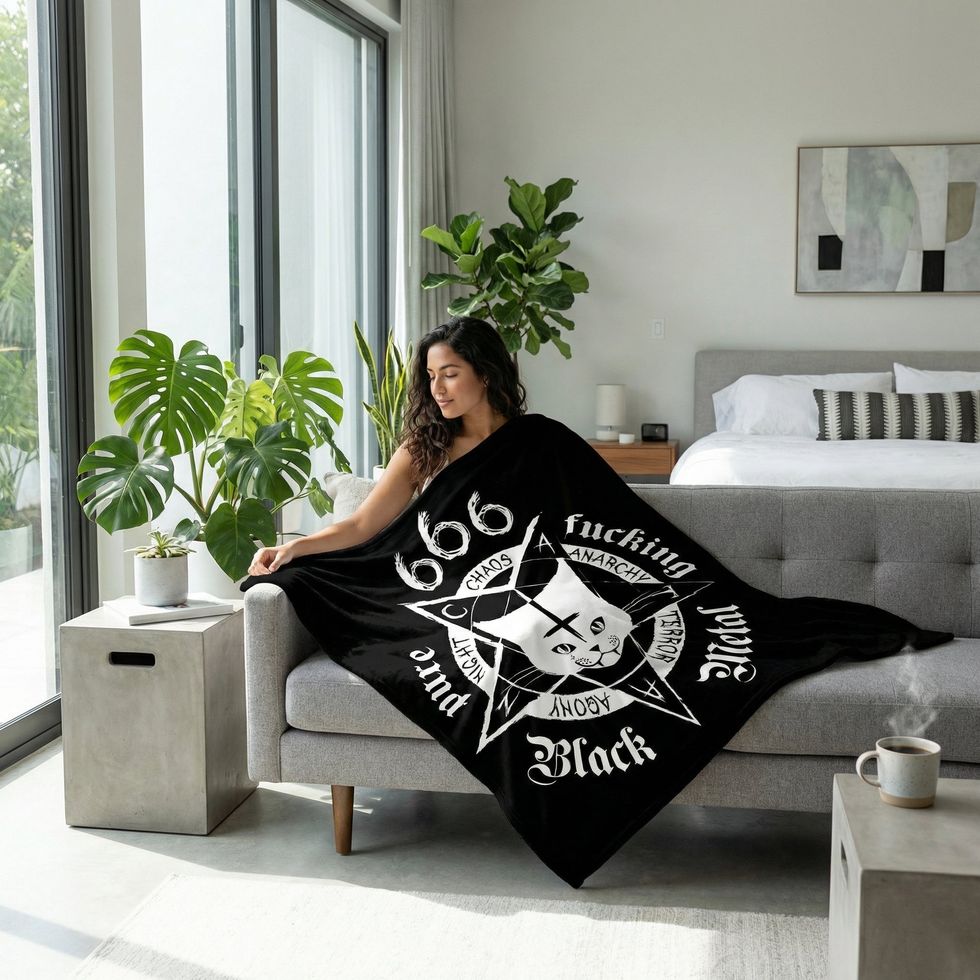 Black flannel throw blanket with a white occult cat and star design, draped over a woman on a gray sofa.