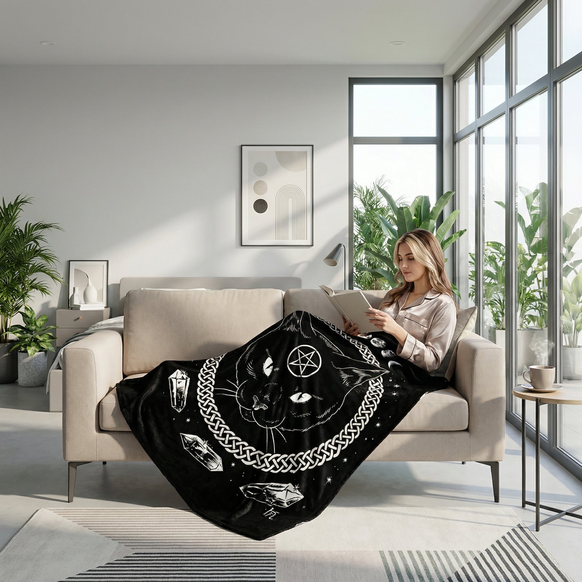 Black flannel throw blanket with a white cat, pentagram, and crystal design, draped over a woman reading on a sofa.