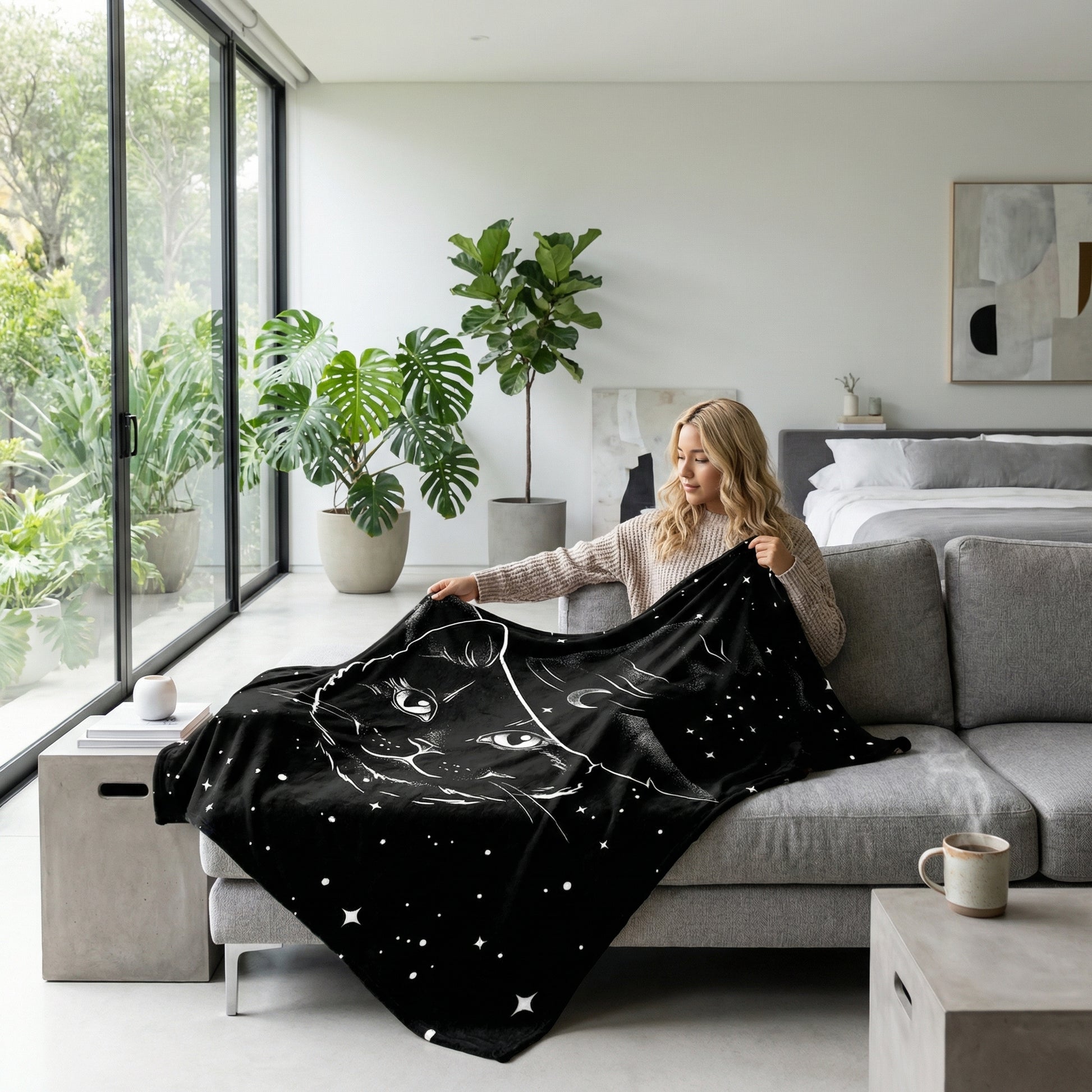 Woman on a gray sofa holding a black flannel throw blanket with a white cat and moon design.