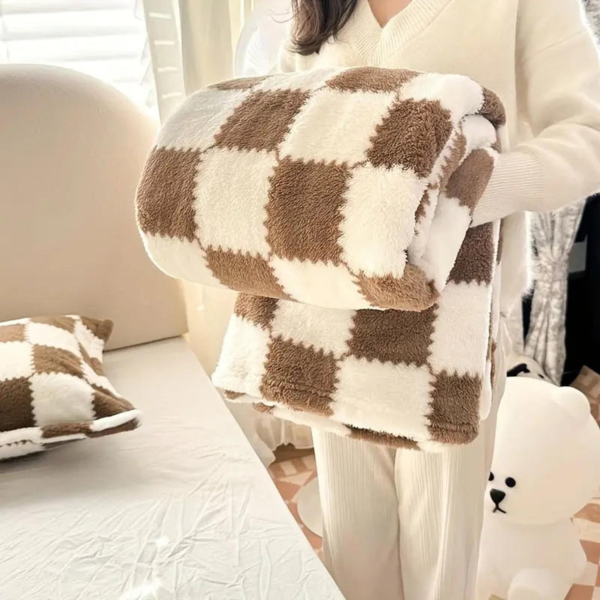 Person holding a folded brown and white checkered flannel fleece blanket with a matching pillow on a bed.