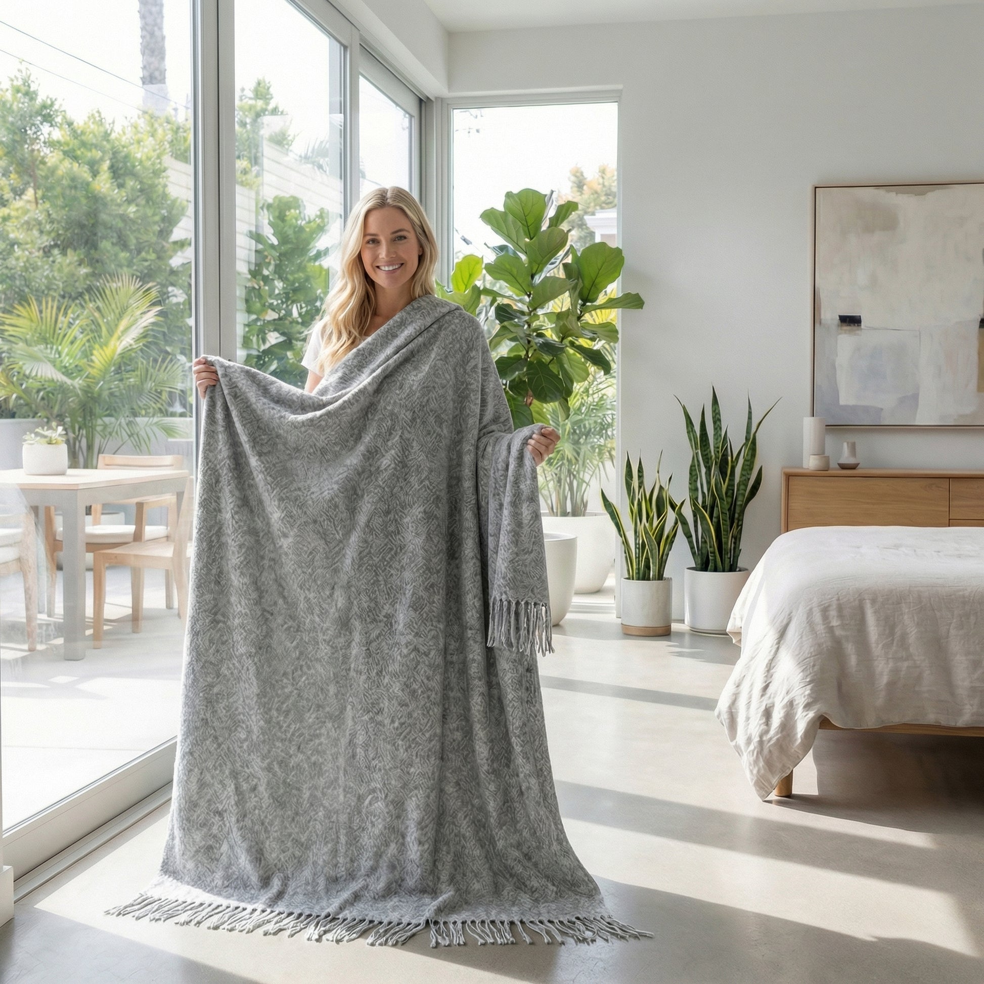 Smiling woman holding a large gray chenille throw blanket with fringes in a bright room.