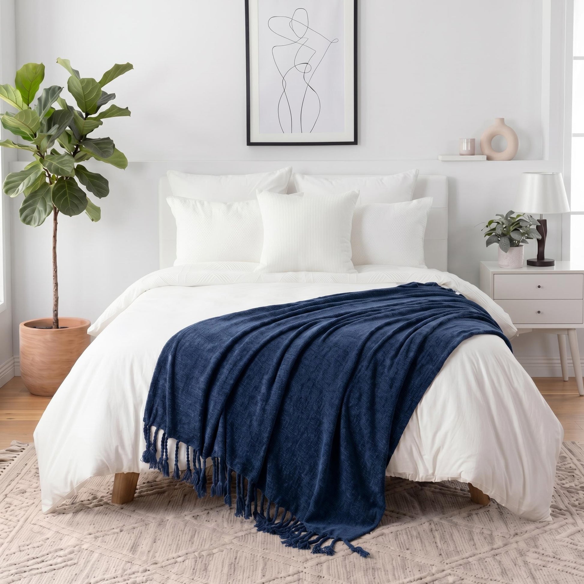 Navy blue chenille throw blanket with fringe tassels draped over a white bed in a bright bedroom.