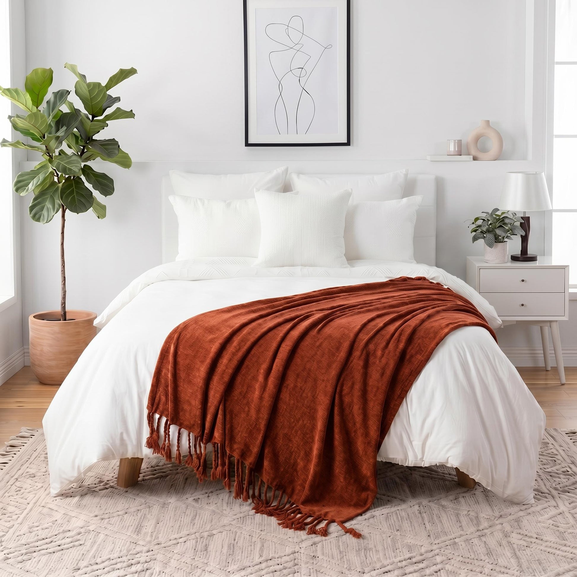 Rust orange chenille throw blanket with fringe tassels draped over a white bed in a bright bedroom.