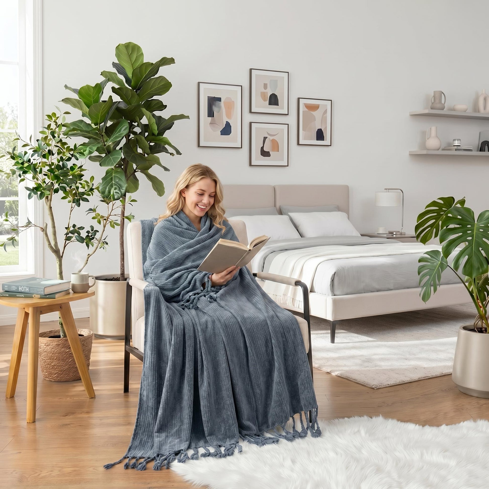 Smiling woman wrapped in a blue chenille throw blanket with fringe tassels, reading in a cozy bedroom.