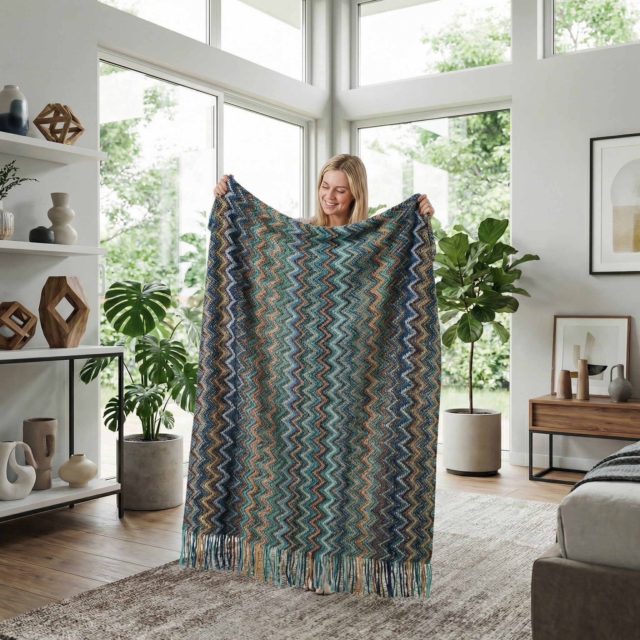 Smiling woman holding a large, hand-knitted blanket with a blue, green, and brown chevron pattern and tassels.