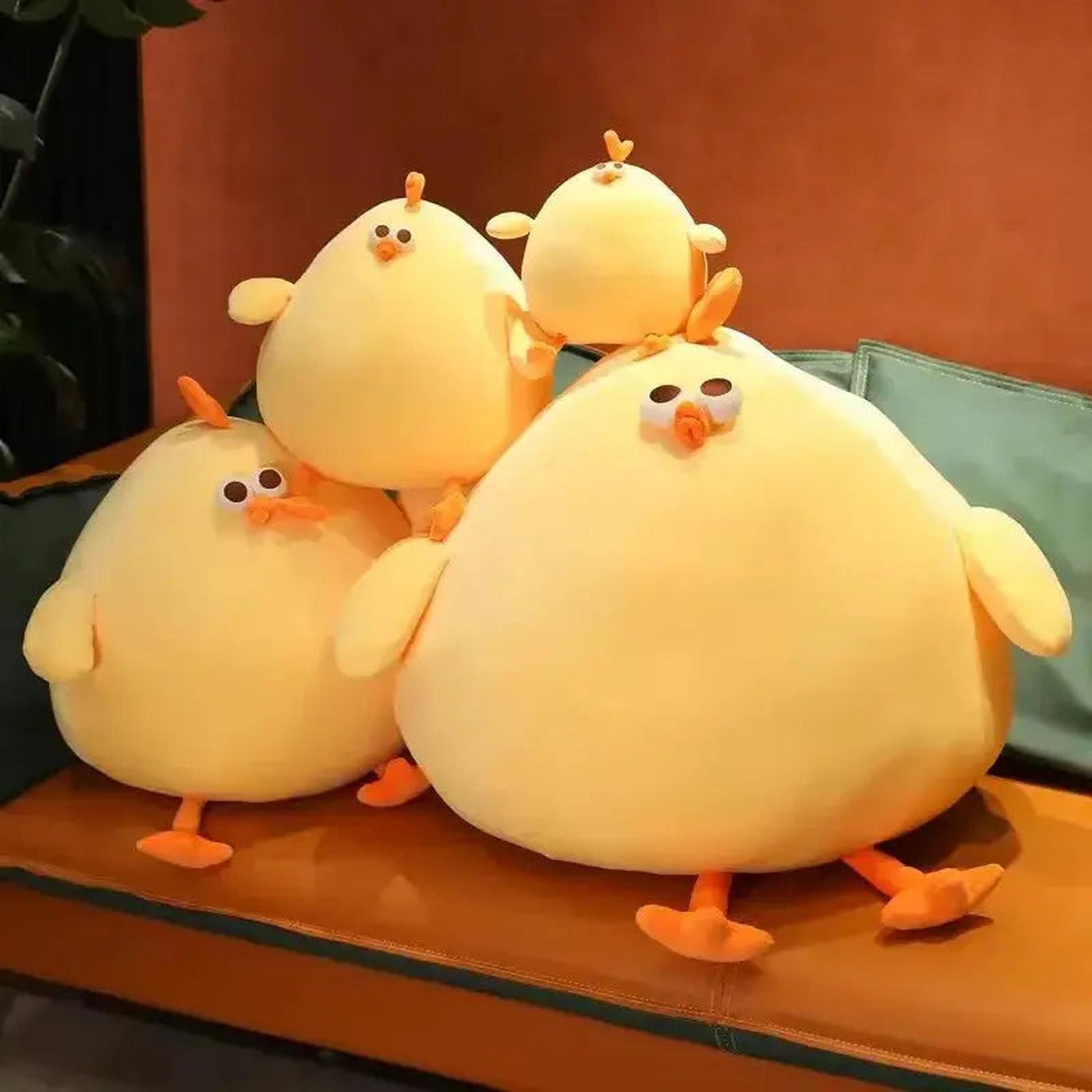 Four bright yellow, round, plush chicken pillows with orange feet and beaks on a brown and green couch.