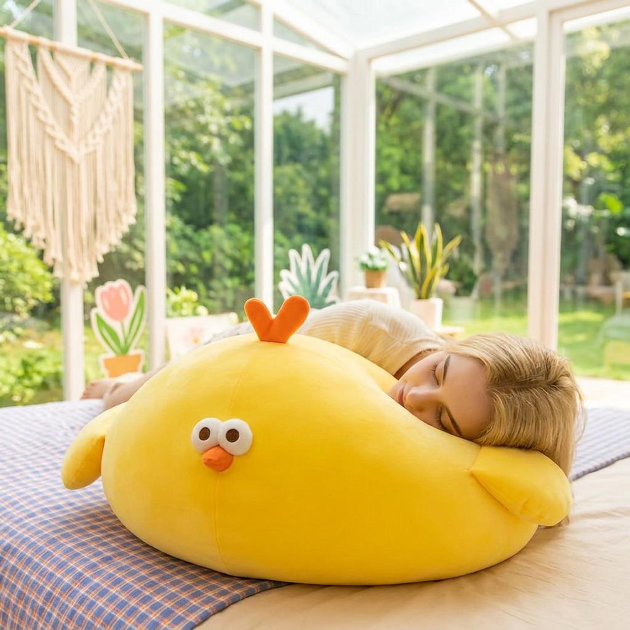 Bright yellow round plush chicken pillow with orange beak and crest, used as a comfortable cushion by a sleeping person.