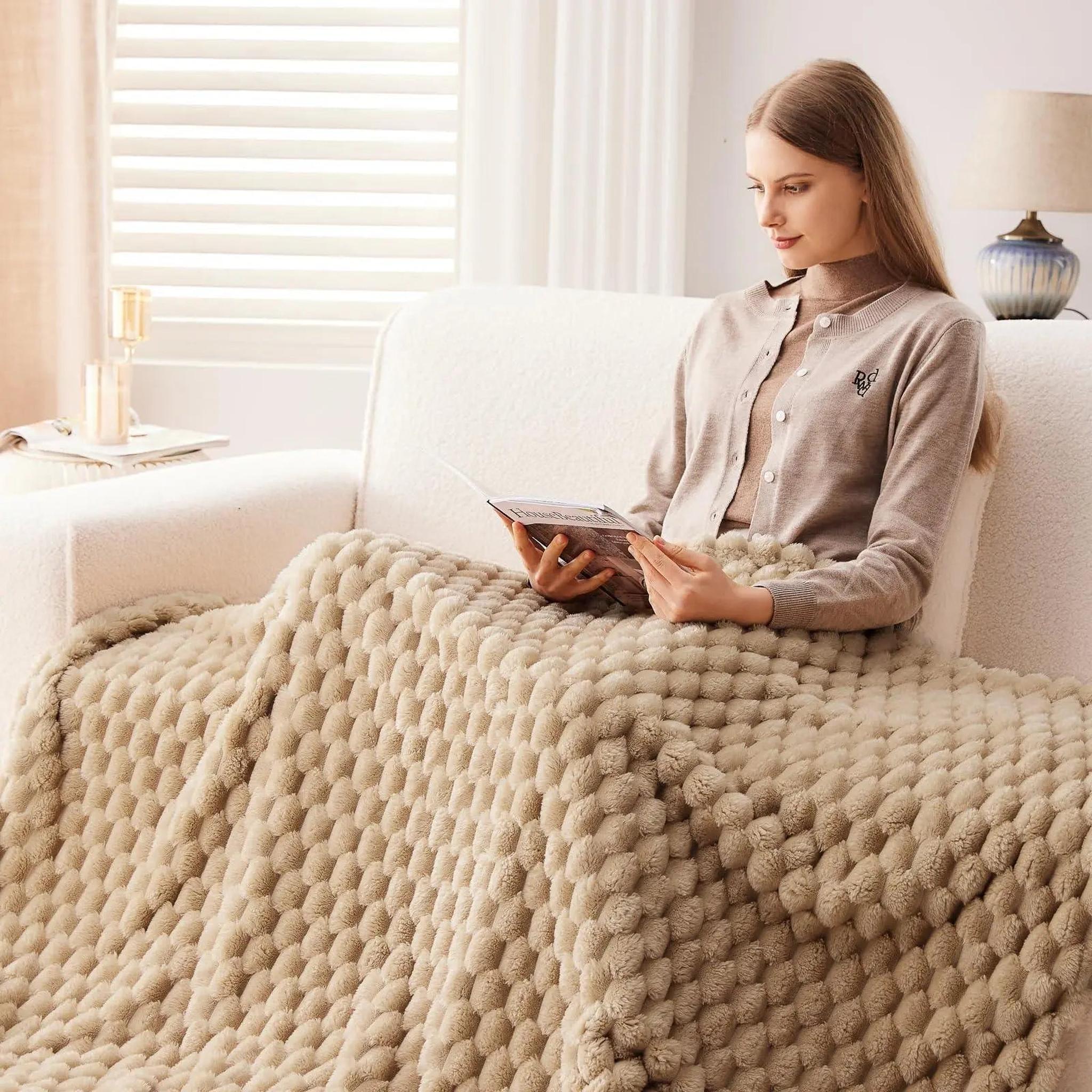 Woman relaxing on a sofa, covered by a beige Clouds Jacquard Fleece Blanket with a 3D cloud pattern.