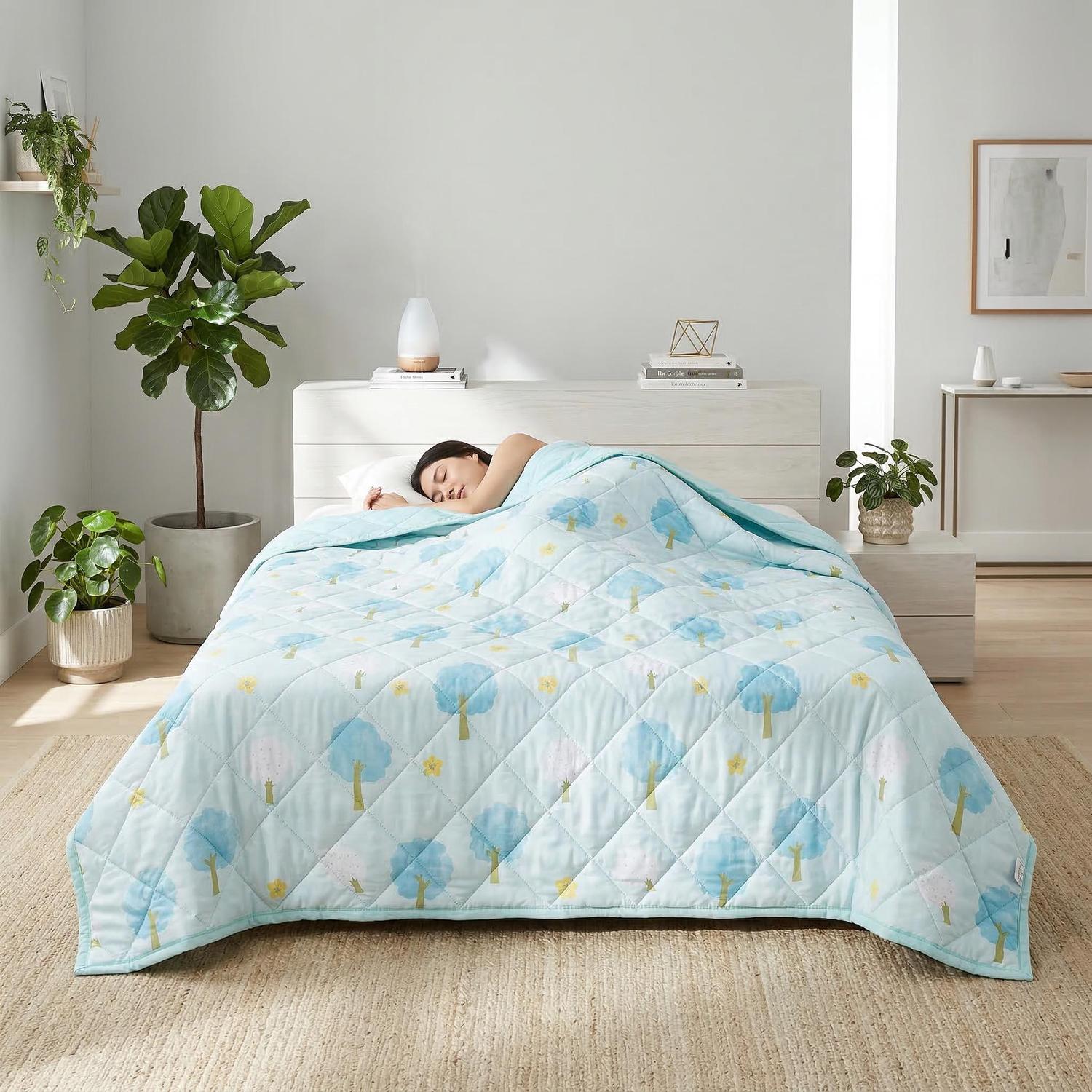 Light blue quilted cooling blanket with a tree pattern covering a sleeping person in a bright bedroom.