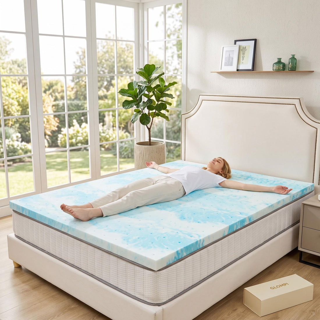 Woman unrolling a thick, blue-marbled cooling gel memory foam mattress topper onto a bed in a bright bedroom.