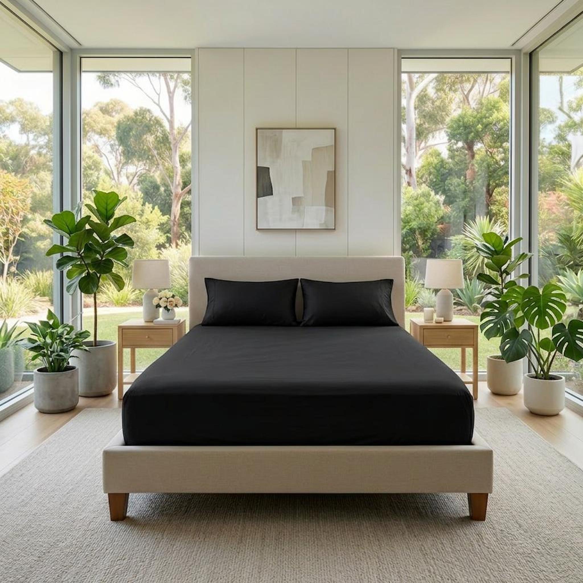 Black cotton fitted sheet on a bed with two pillows, in a bright bedroom with large windows.