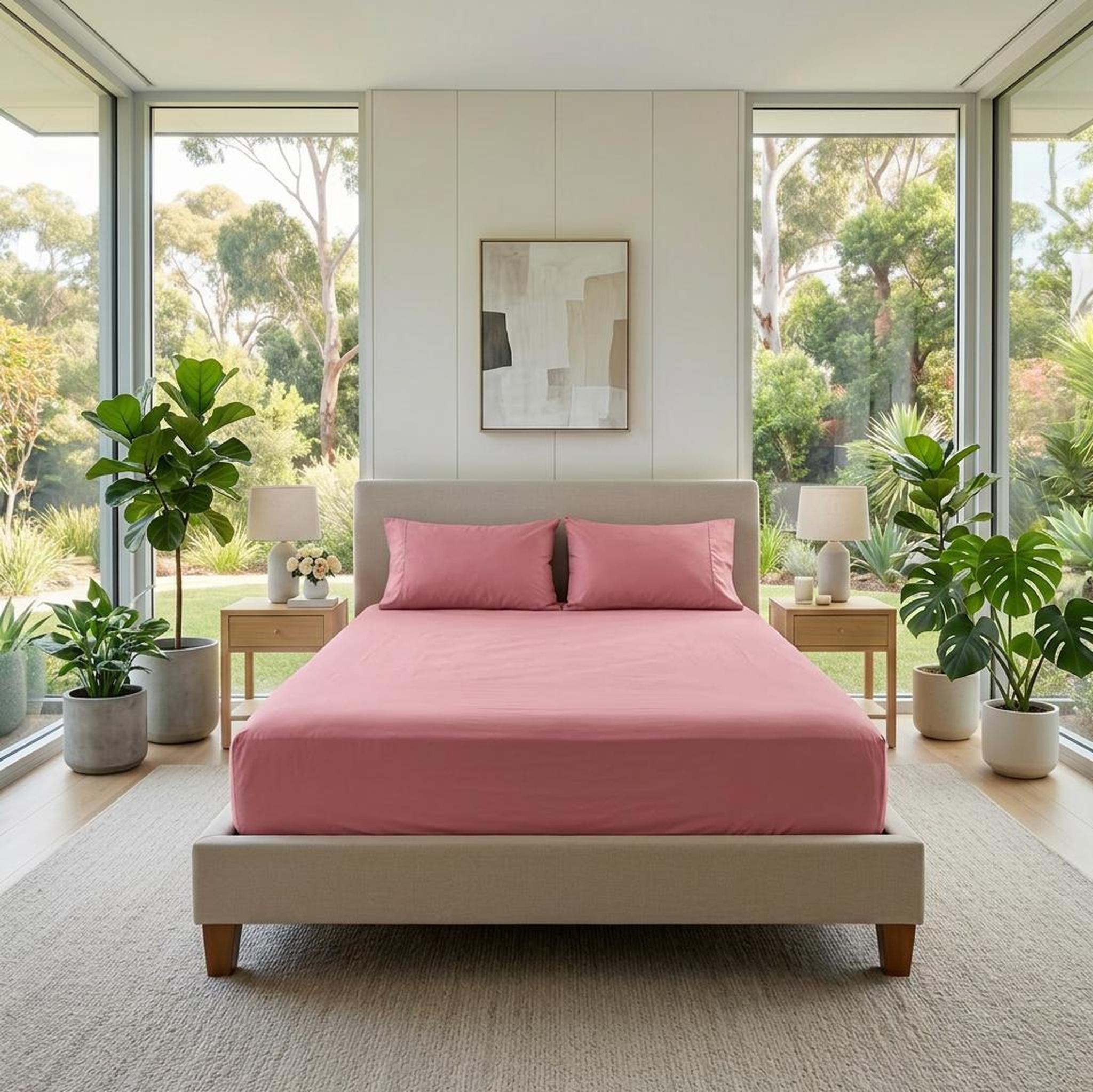 Pink cotton fitted sheet and pillowcases on a bed in a bright bedroom with large windows.