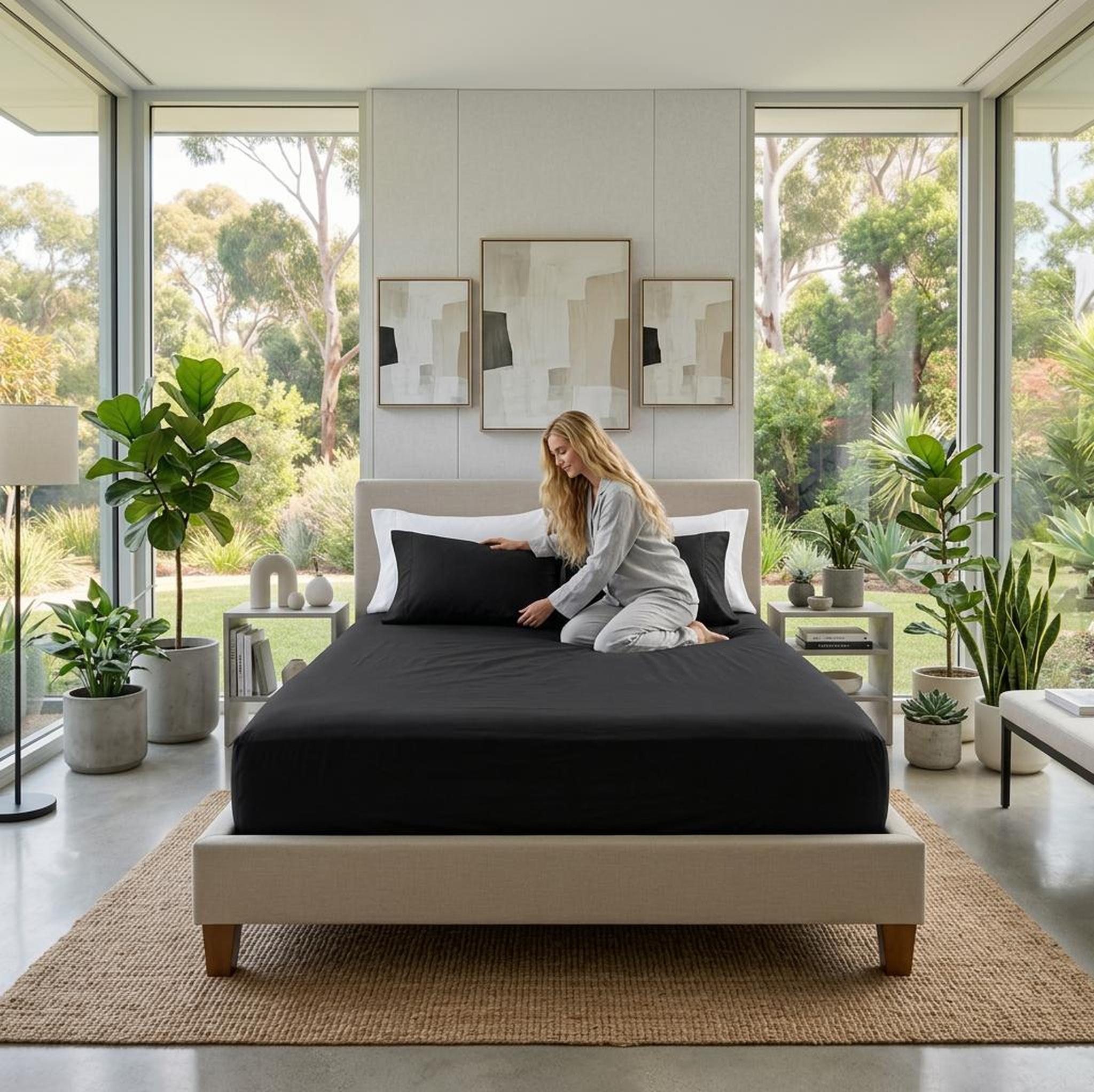 Woman on a bed with a black cotton fitted sheet and pillowcases, in a modern bedroom with large windows.